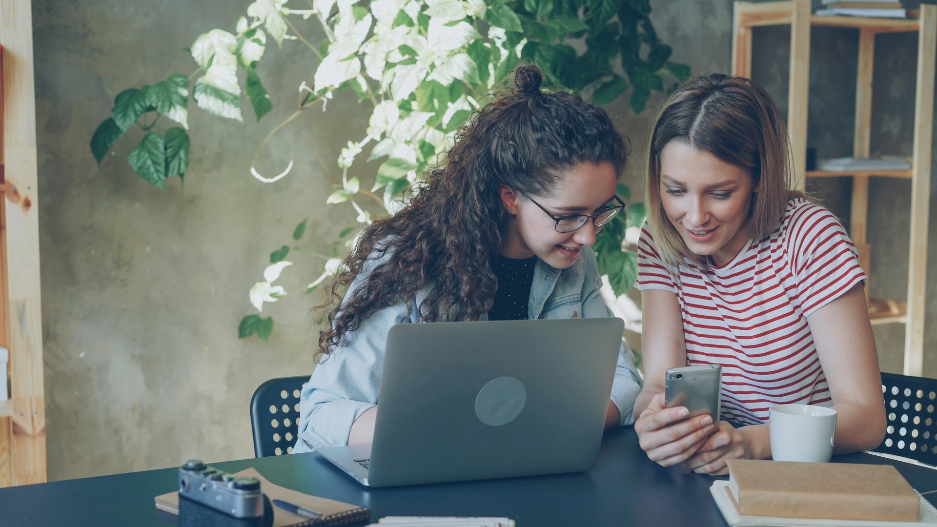 Two women look at a phone next to a laptop.