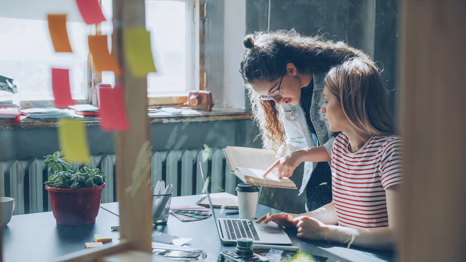 Two women collaborate over a laptop in an office, discussing a project together.