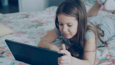 A girl is using a tablet on her bed.