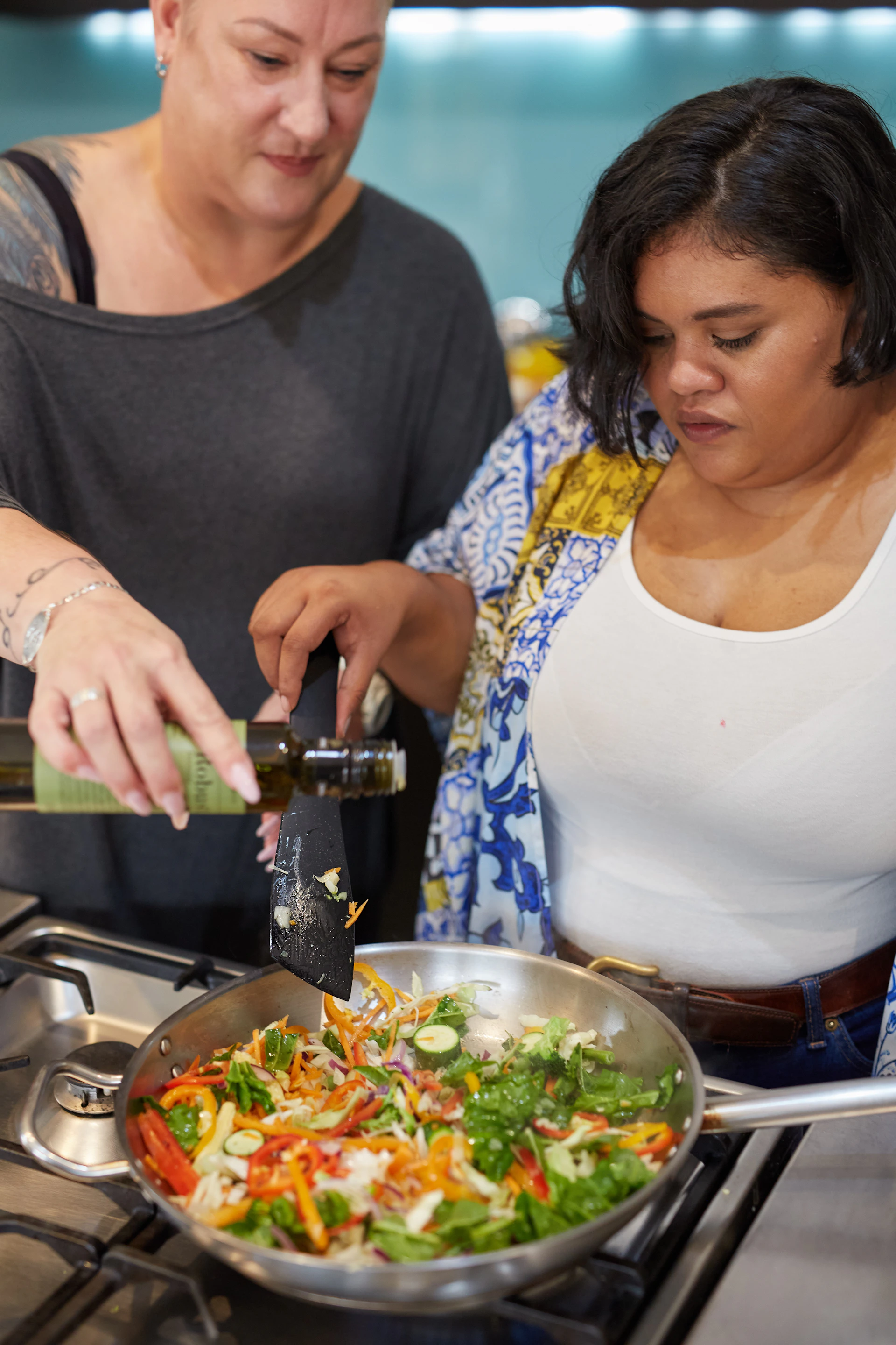 Two women cook vegetables together in a pan.
