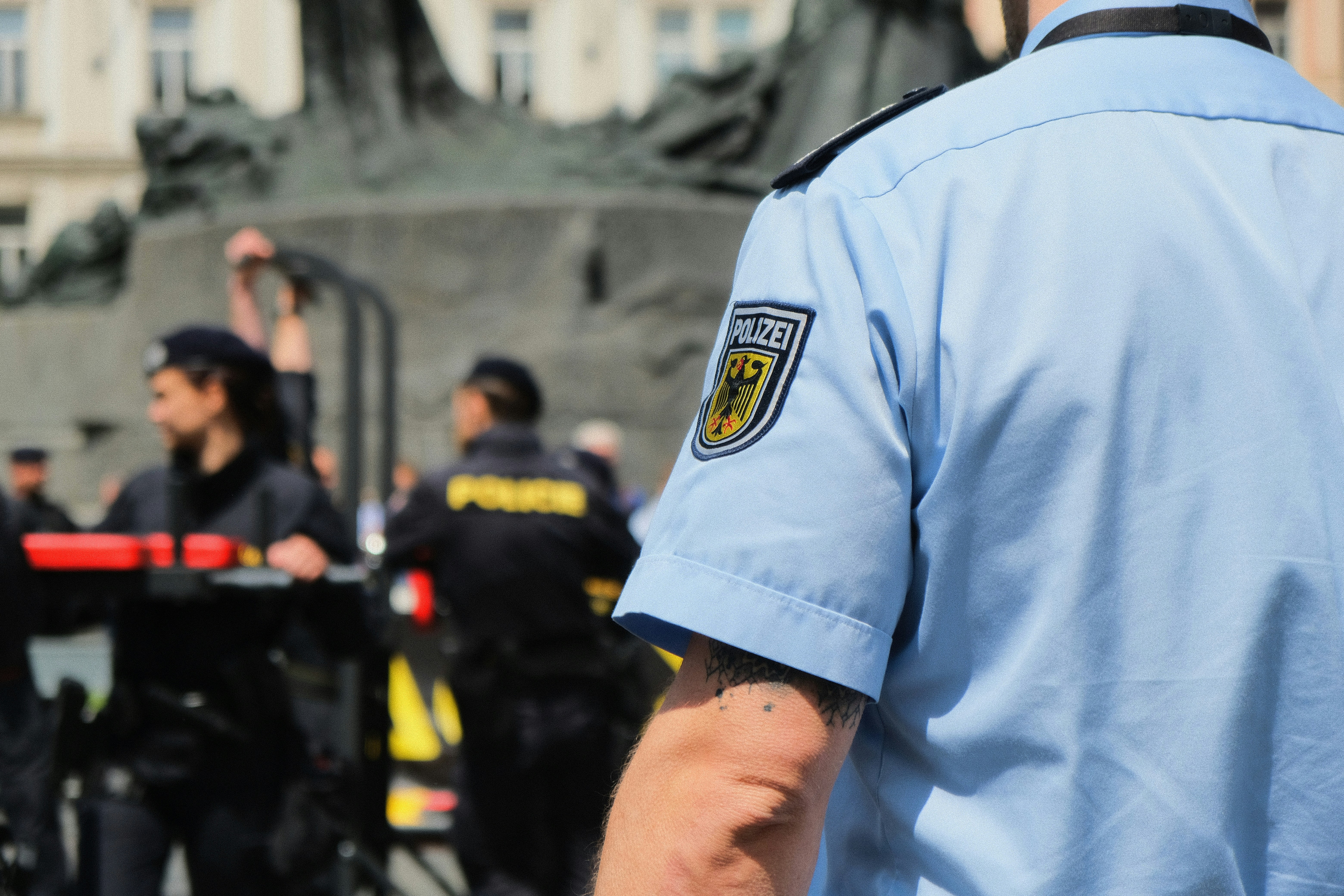 German police officer in uniform seen from behind, with other officers managing a public area in the background.