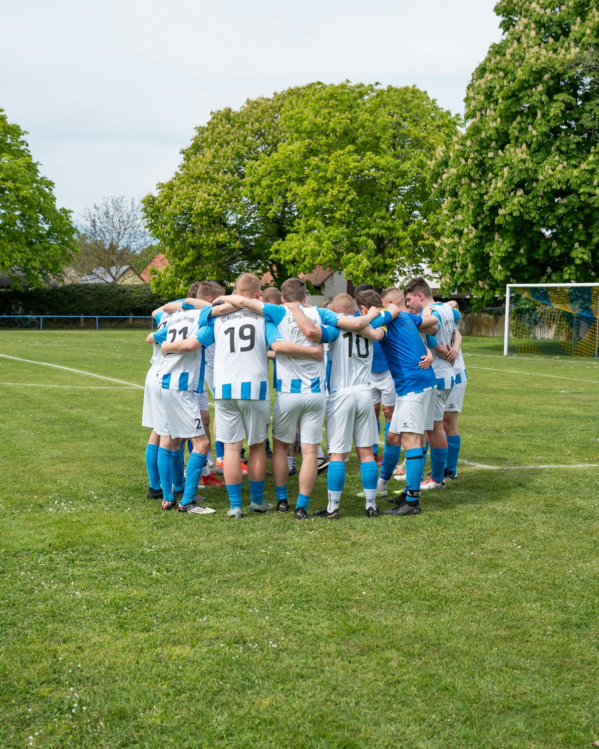 Équipe de football amateur en maillots rayés blanc et bleu, regroupée en cercle sur un terrain en herbe bordé d'arbres