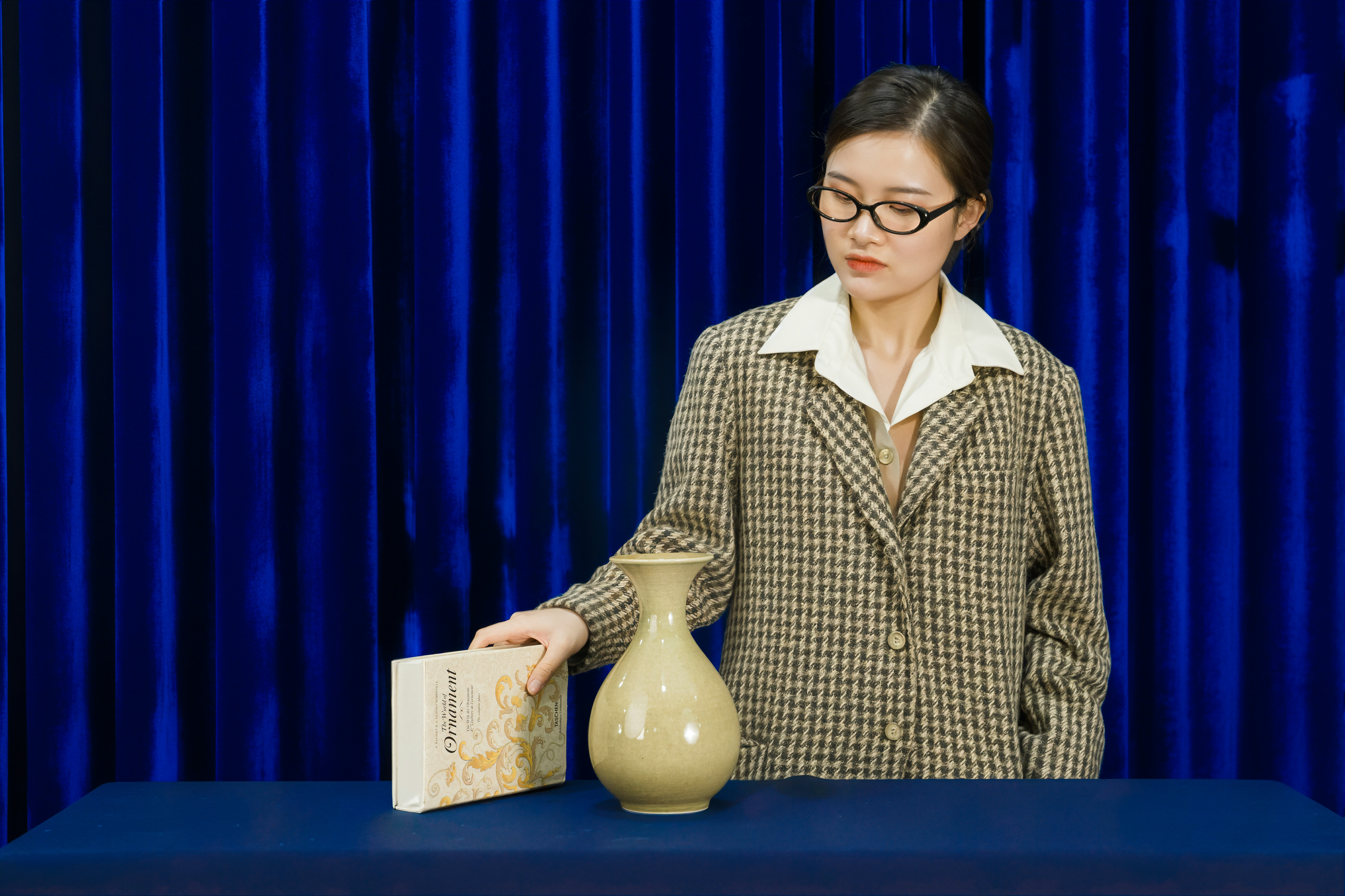 Woman in blazer examines vase and book.