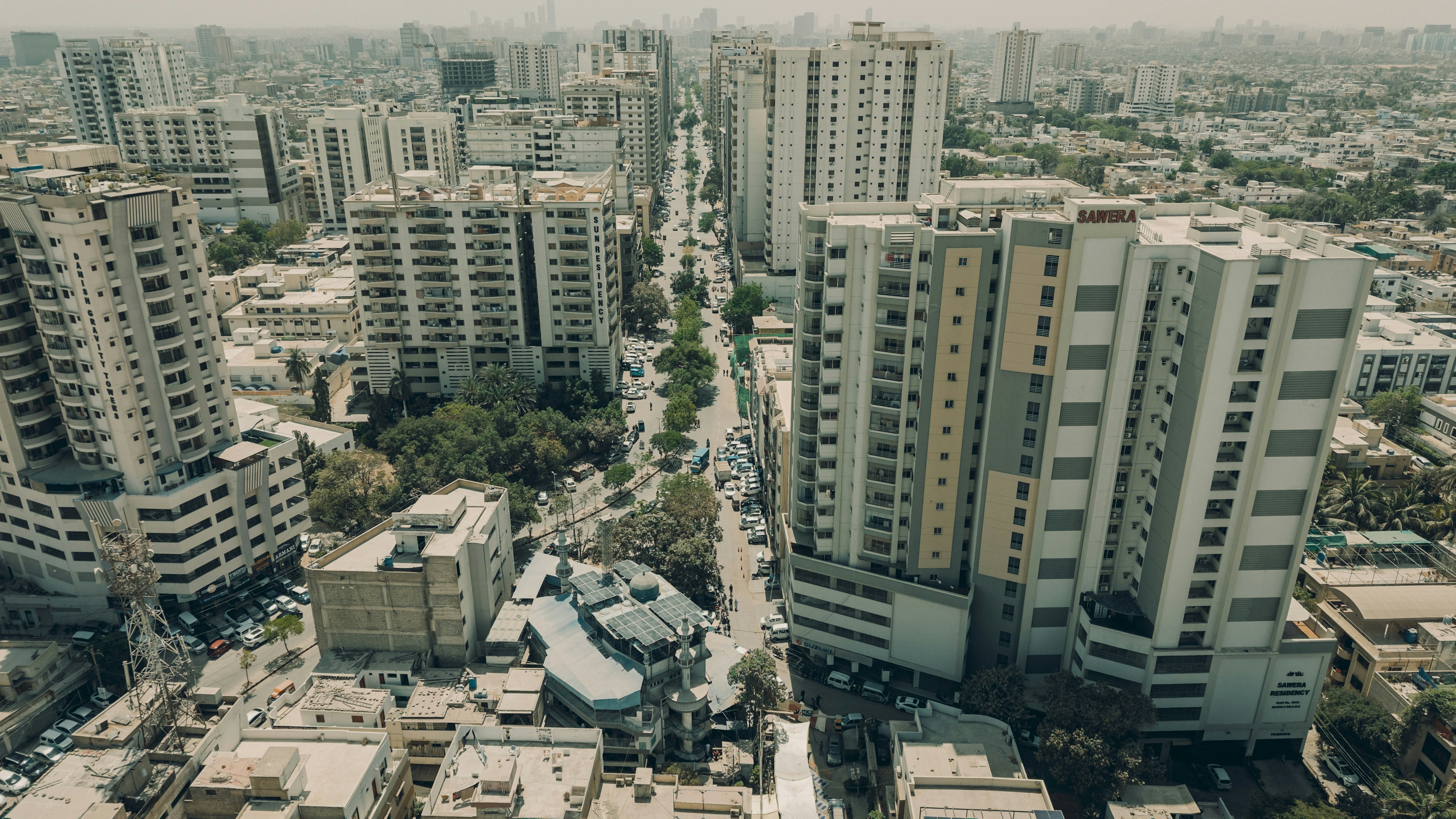 A cinematic aerial photograph capturing a symmetrical urban street in Karachi, Pakistan, flanked by tall residential and commercial buildings. The shot highlights the dense infrastructure, traffic movement, and vertical planning of the city’s modern landscape. Taken during daylight with a clean and moody tone for editorial or documentary use.