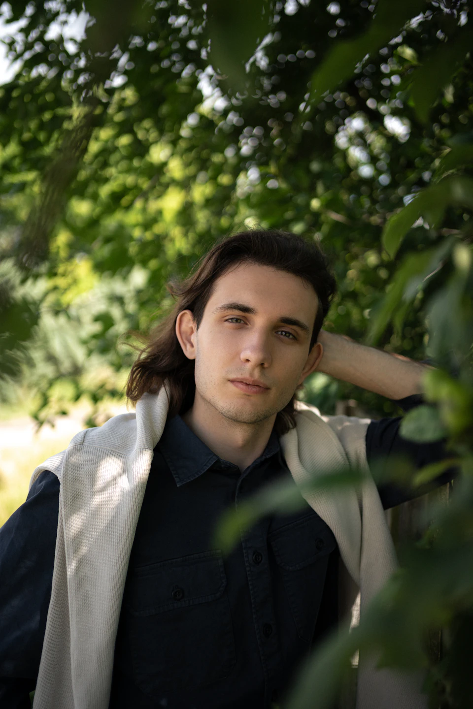 In the image, a young man with long hair is captured leaning against a tree trunk and looking directly at the camera. He's dressed casually in a blue shirt paired with a white jacket. The background features lush green leaves from trees, providing a natural backdrop to the scene.