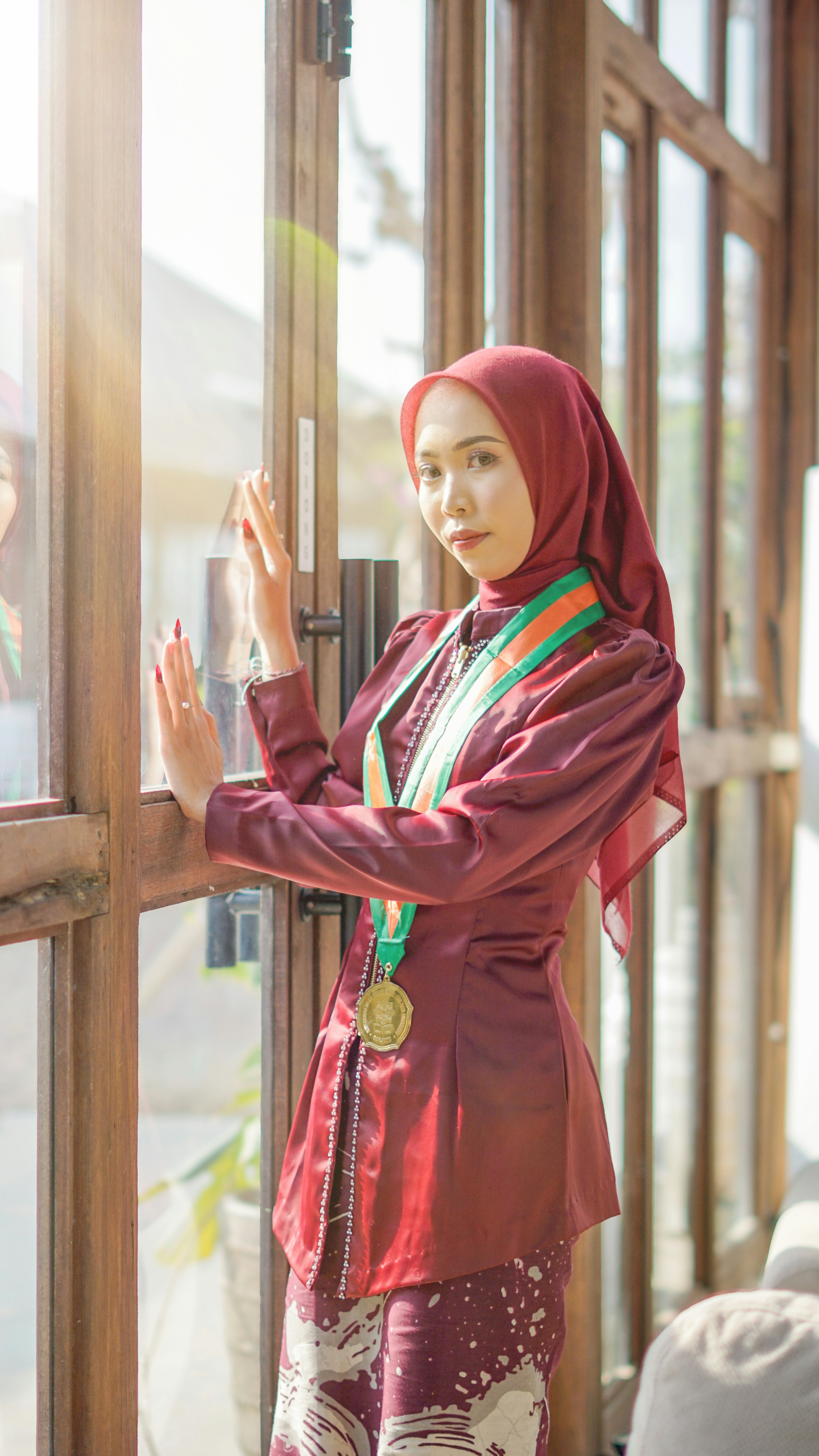 A woman poses by a window wearing graduation attire.