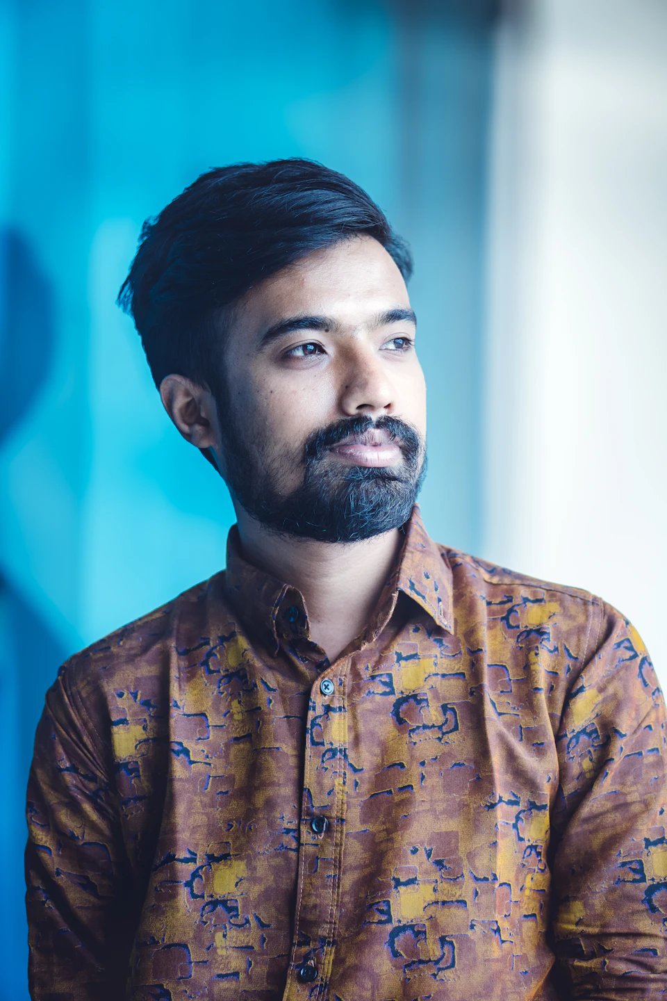 The image shows a man with dark hair and a beard wearing a brown shirt adorned with blue and yellow floral patterns, standing against a light blue background. The man's gaze is directed off to the side, suggesting he may be lost in thought or observing something outside of the frame.