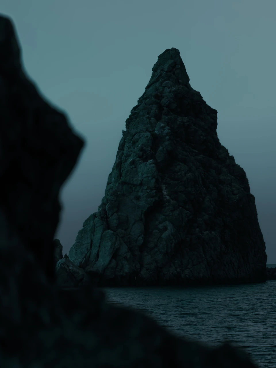 Dark green rock formation with steep top stands out against ocean backdrop on island