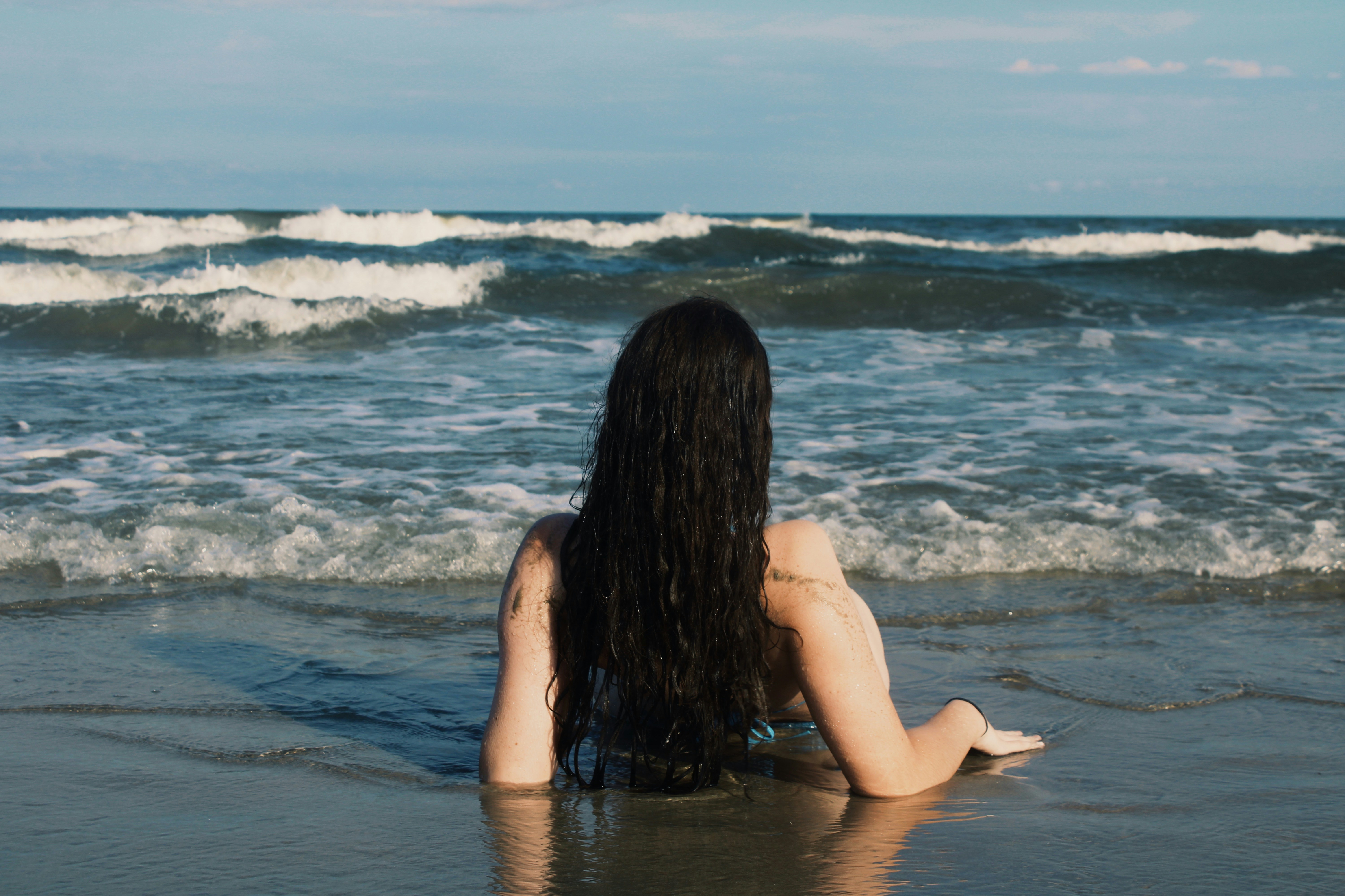 Woman sits in the water, facing the ocean.