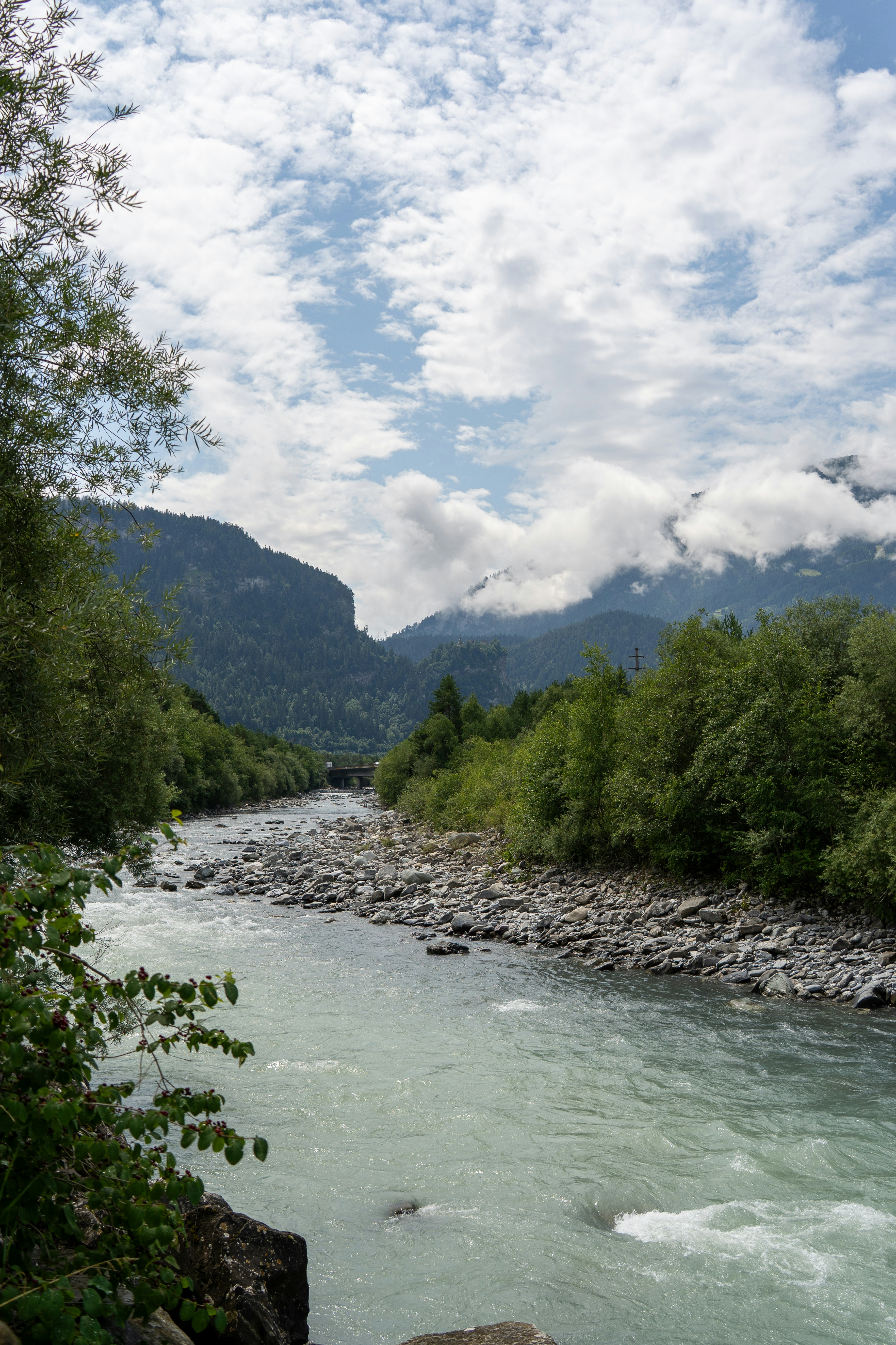 The glacial-blue waters of the Hinterrhein river rush through a rocky alpine valley, framed by dense green forests and mist-covered mountain peaks. Under a patchwork sky, this scene captures the wild, unspoiled beauty of Switzerland’s mountain rivers.