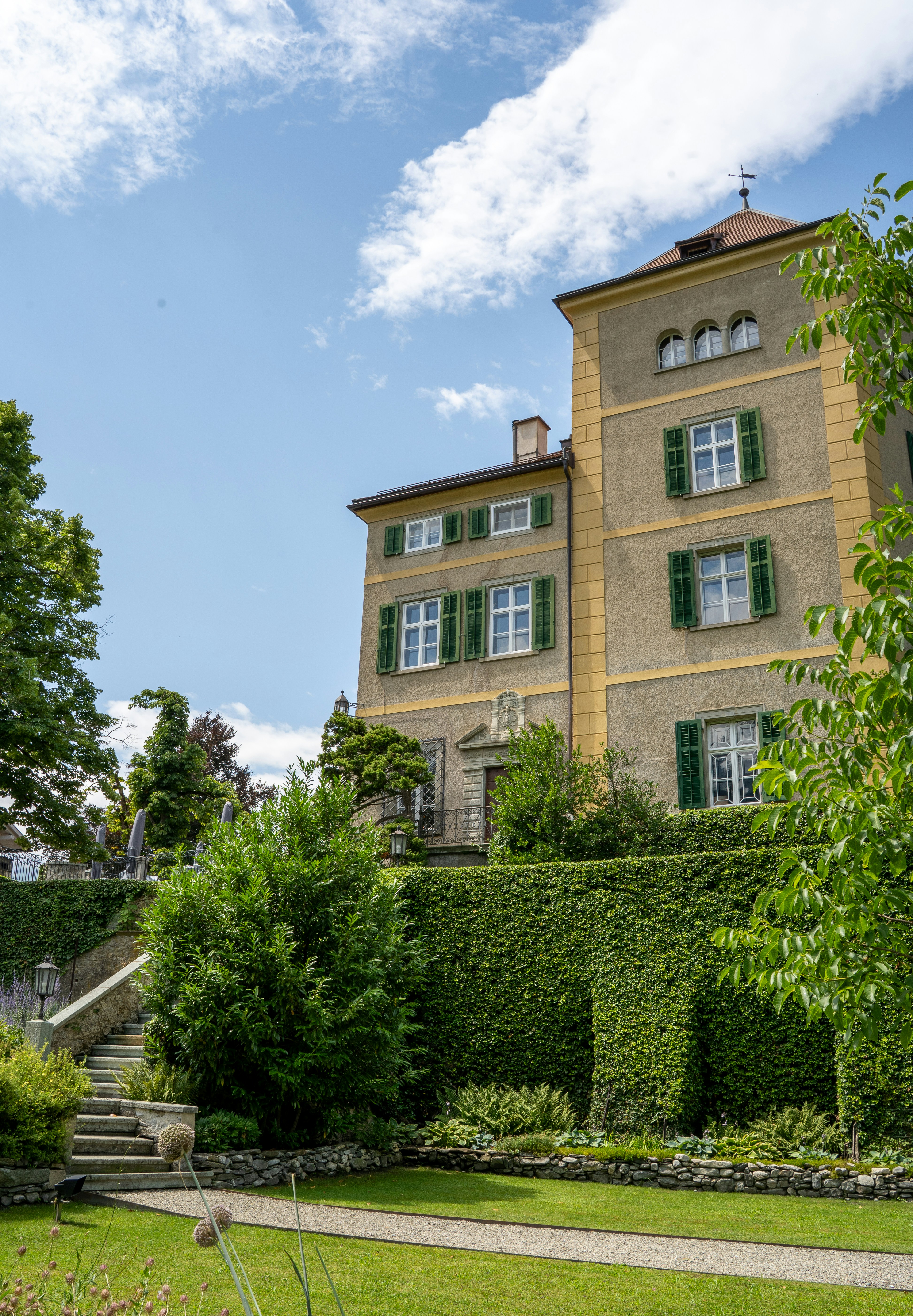 An elegant historic villa rises above manicured gardens and ivy-covered walls, its ochre-toned facade and green shutters glowing under the Swiss summer sun. This peaceful estate evokes timeless charm and refined Alpine architecture.