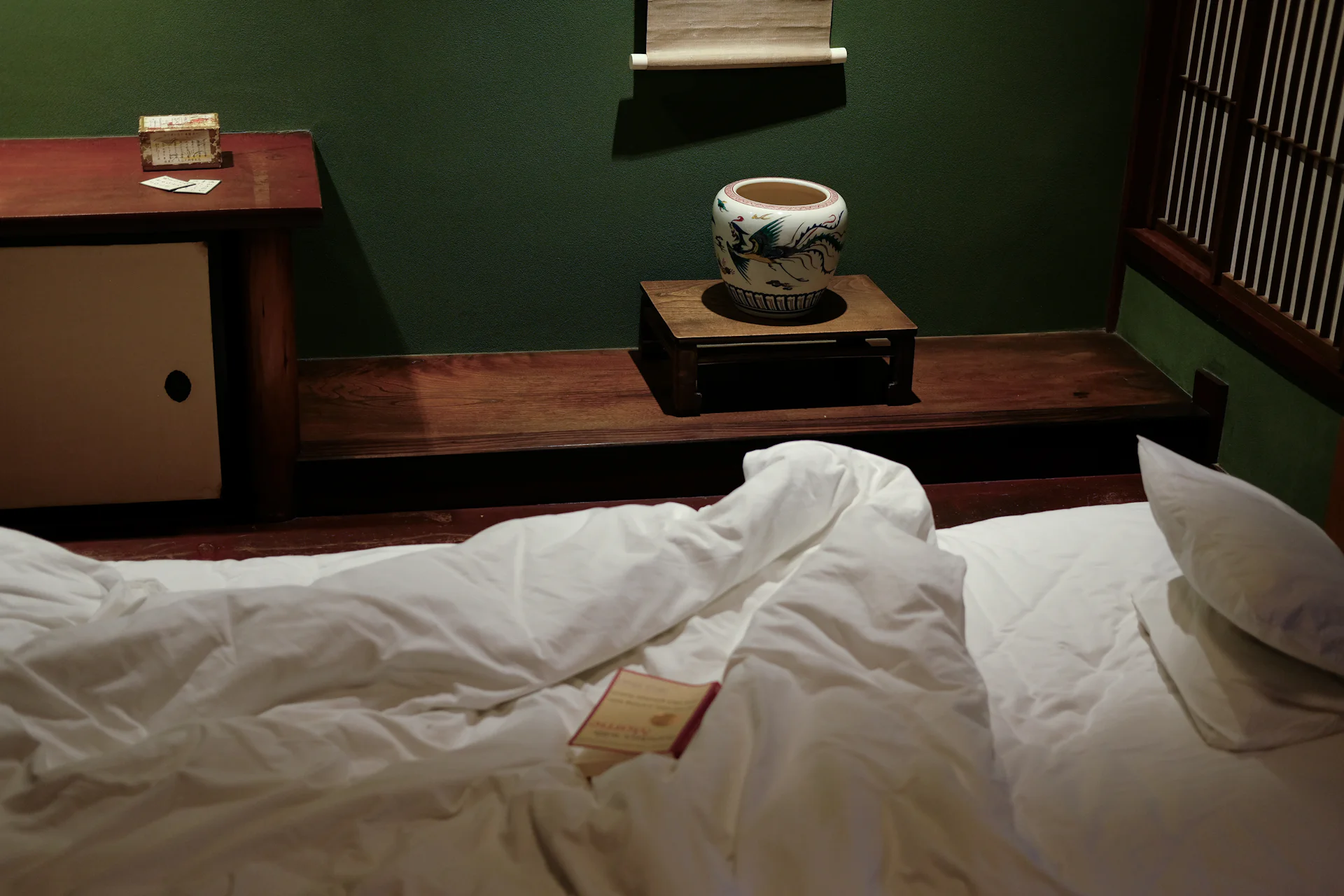 Futon with white bedding in a dim tatami room with shoji and a ceramic jar in the alcove