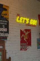 A neon sign says, "let's go!" on a brick wall.