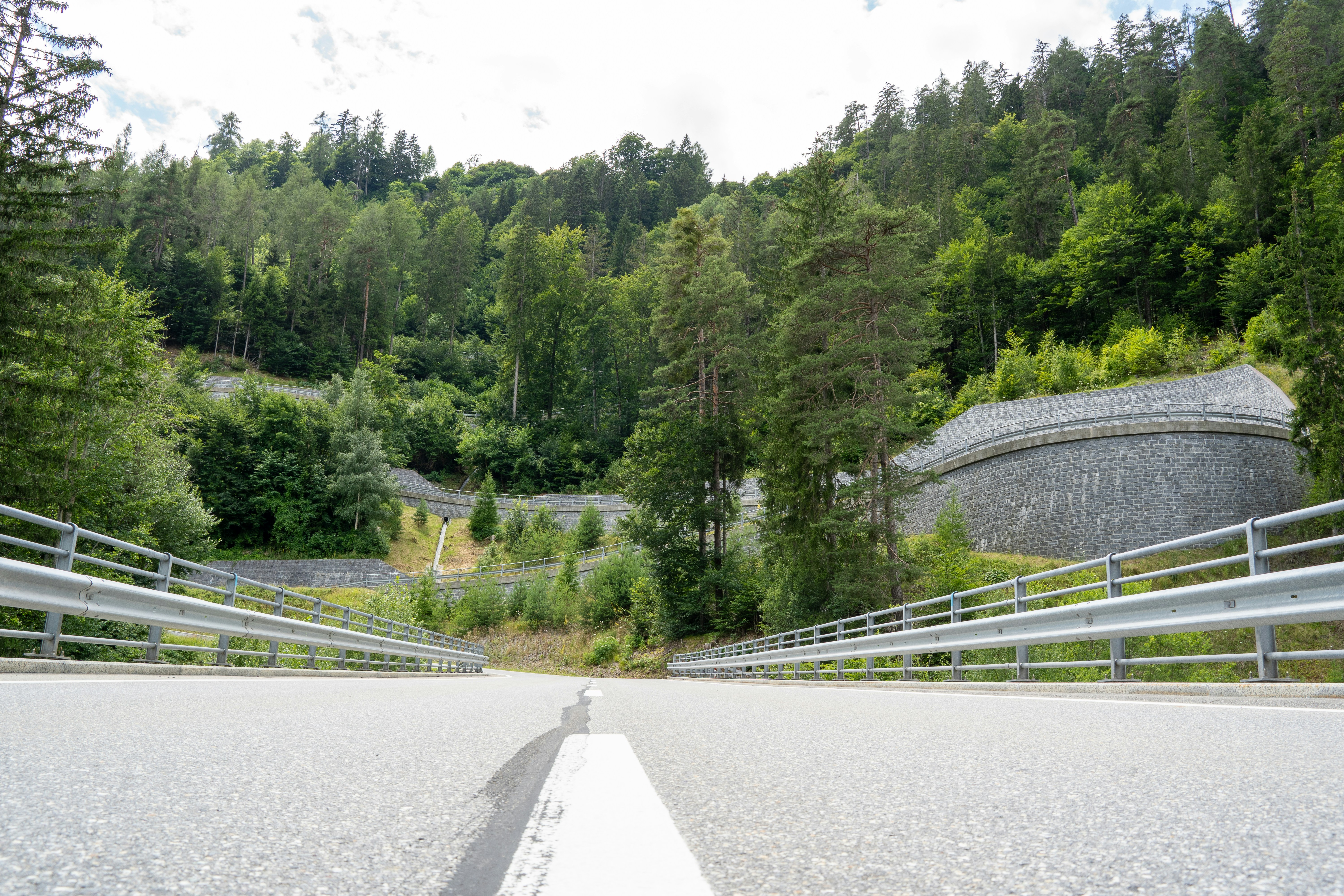 A winding mountain road in Graubünden, Switzerland, cuts through dense forest and climbs along carefully engineered stone retaining walls. Viewed from ground level, the scene blends smooth asphalt, lush greenery, and structured curves, capturing the balance between nature and infrastructure in alpine terrain.