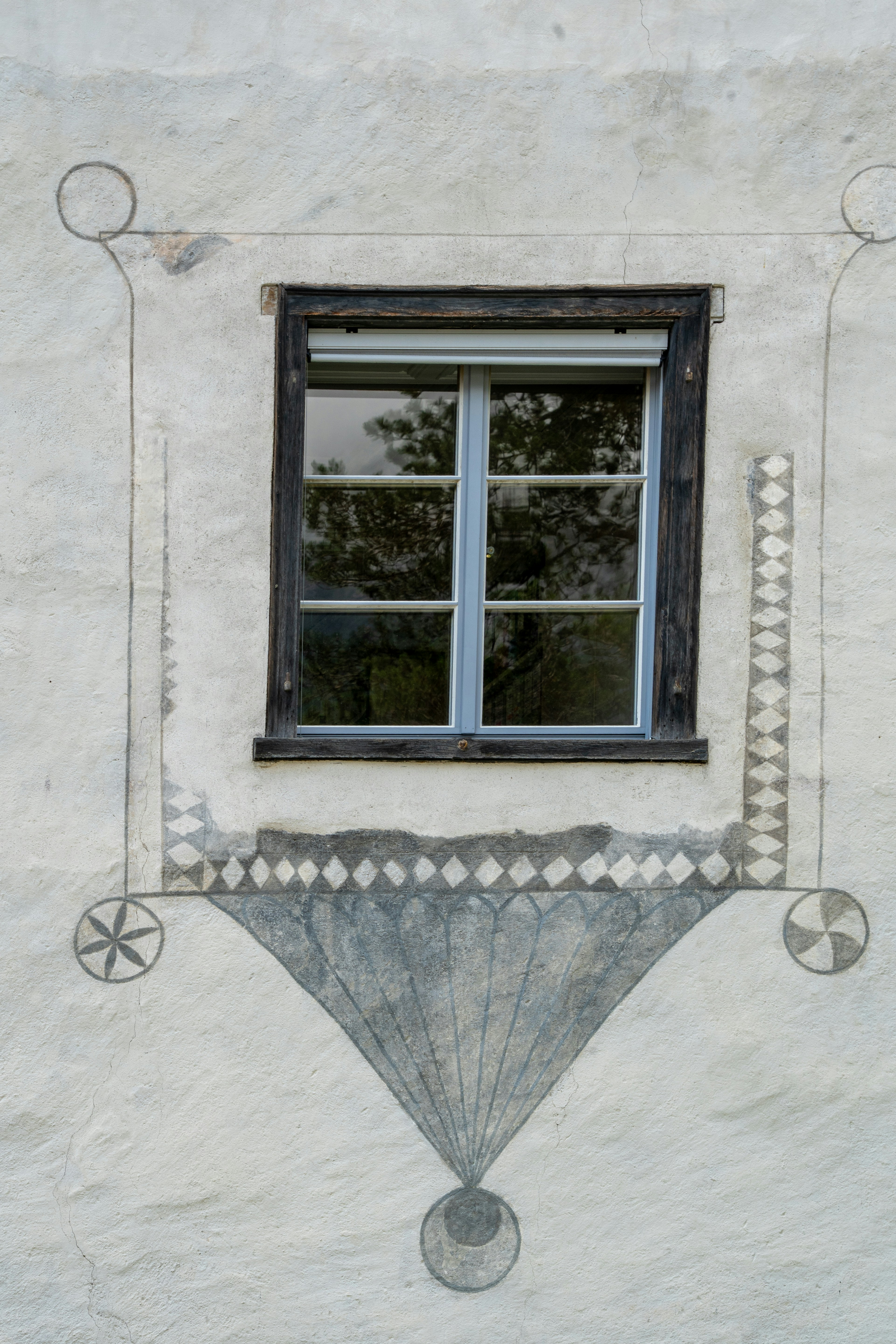 A close-up of a window framed by intricate sgraffito artwork on a historic building in Fürstenau, Switzerland. The geometric patterns and faded detailing add a timeless elegance to the weathered plaster wall, reflecting centuries of Alpine craftsmanship.