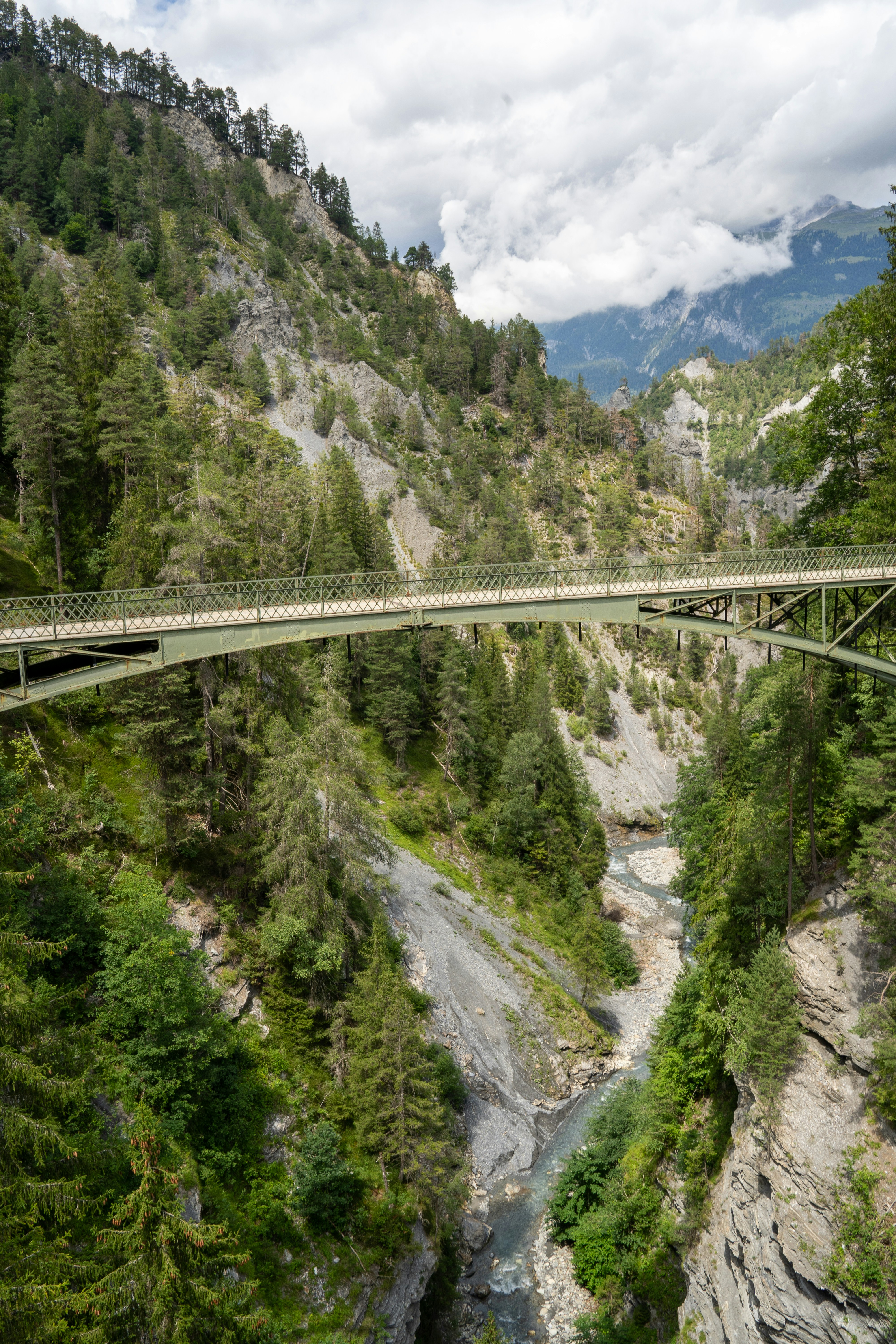 A narrow iron bridge stretches boldly across a deep alpine gorge in Graubünden, Switzerland, surrounded by steep cliffs, pine forests, and a winding river far below. The scene captures the harmony between human engineering and untamed mountain wilderness under a moody, cloud-streaked sky.