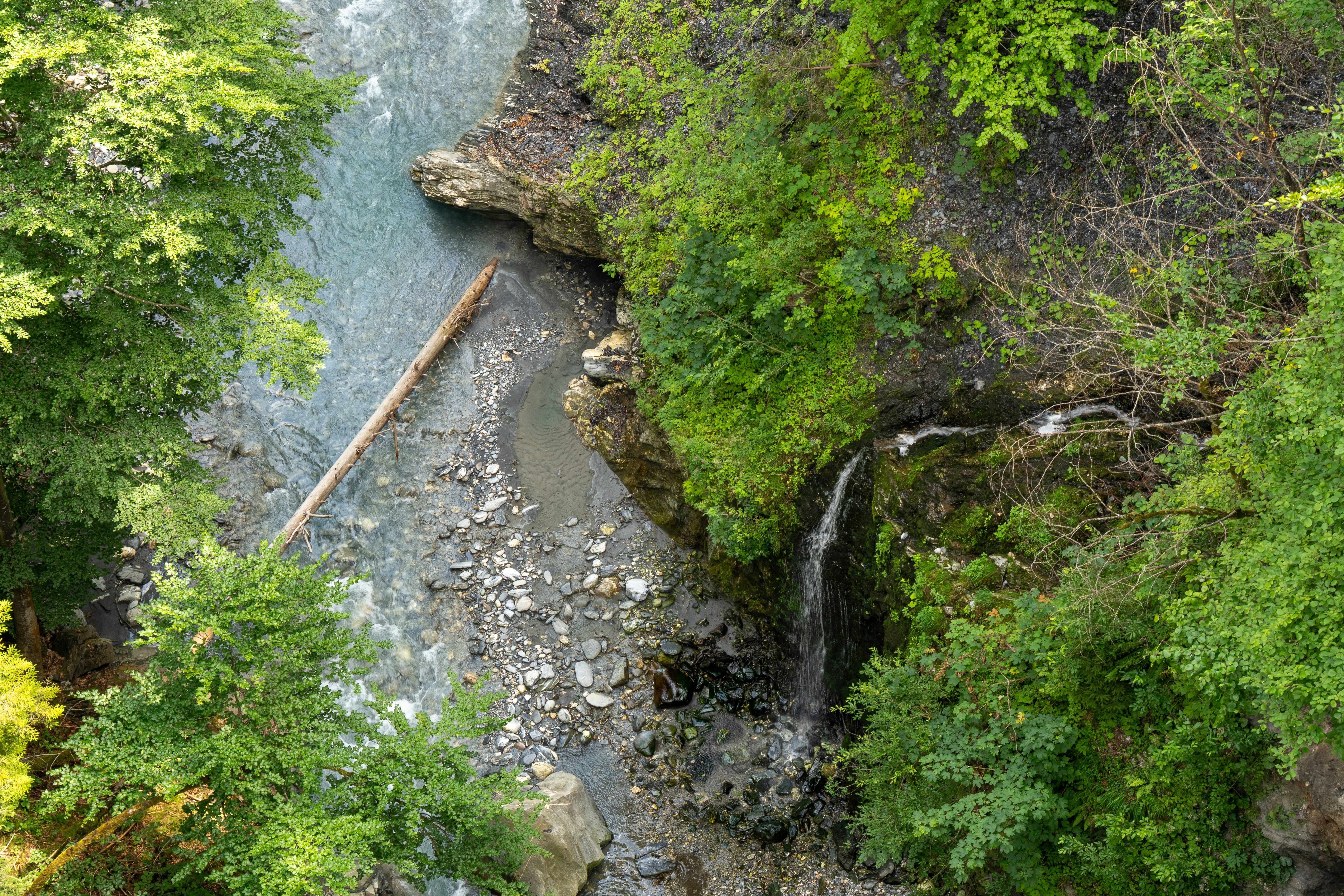 A small alpine waterfall spills gently over moss-covered rock into a stony mountain stream in Graubünden, Switzerland. Surrounded by lush green foliage and the scattered remnants of driftwood, the scene captures a quiet, untouched corner of the wilderness.
