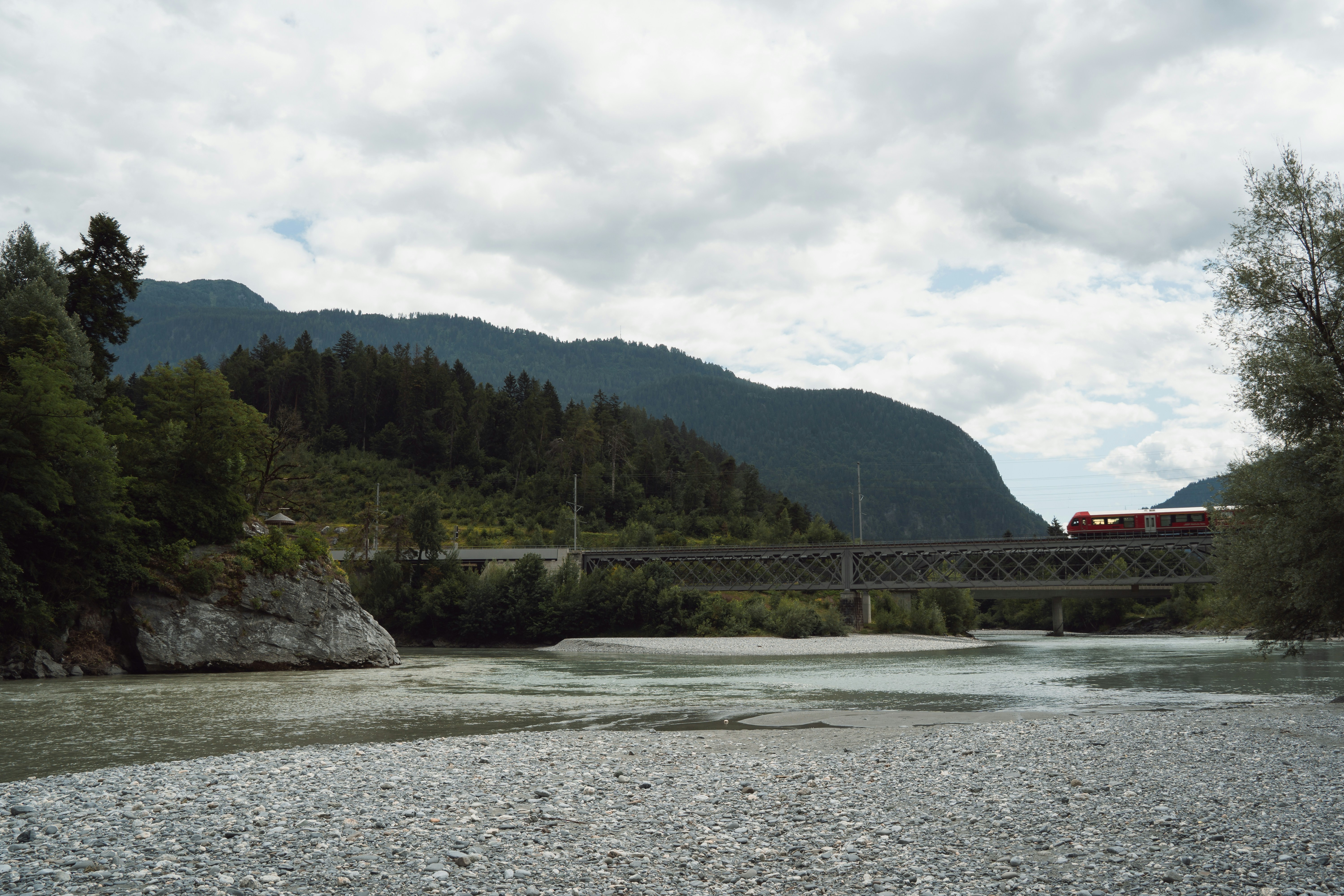 At the confluence of the Hinterrhein and Vorderrhein rivers in Switzerland, a red train crosses a steel bridge framed by forested hills and alpine peaks. The gravel riverbank and gentle current create a peaceful scene where nature and movement intersect.