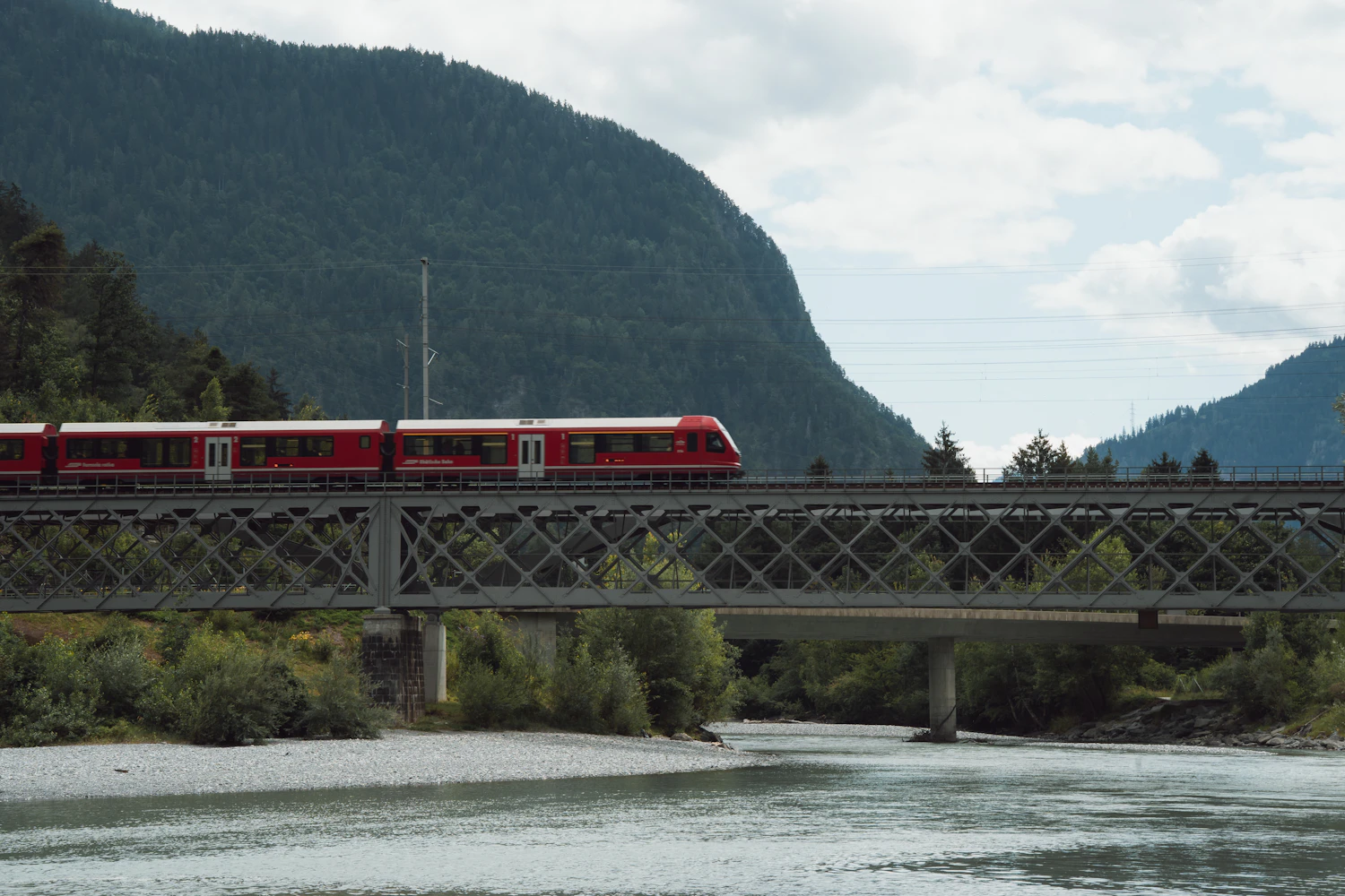 Tren rojo cruzando un puente sobre un río entre montañas de los Alpes suizos, Suiza