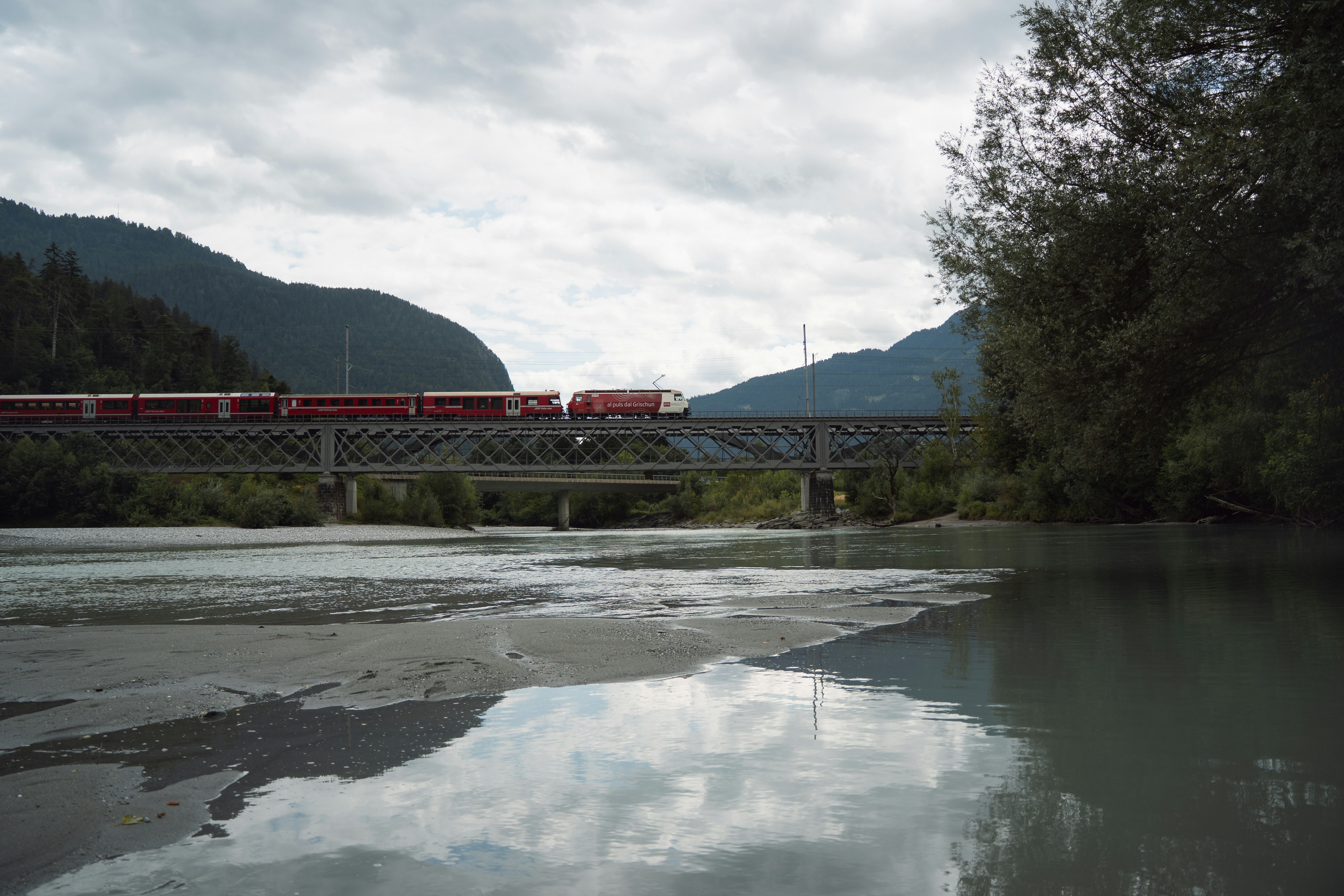 A red Swiss train crosses a steel bridge over the calm, reflective waters at the confluence of the Hinterrhein and Vorderrhein. With moody clouds above and tranquil mountain scenery all around, the image captures the quiet rhythm of travel through the heart of nature.