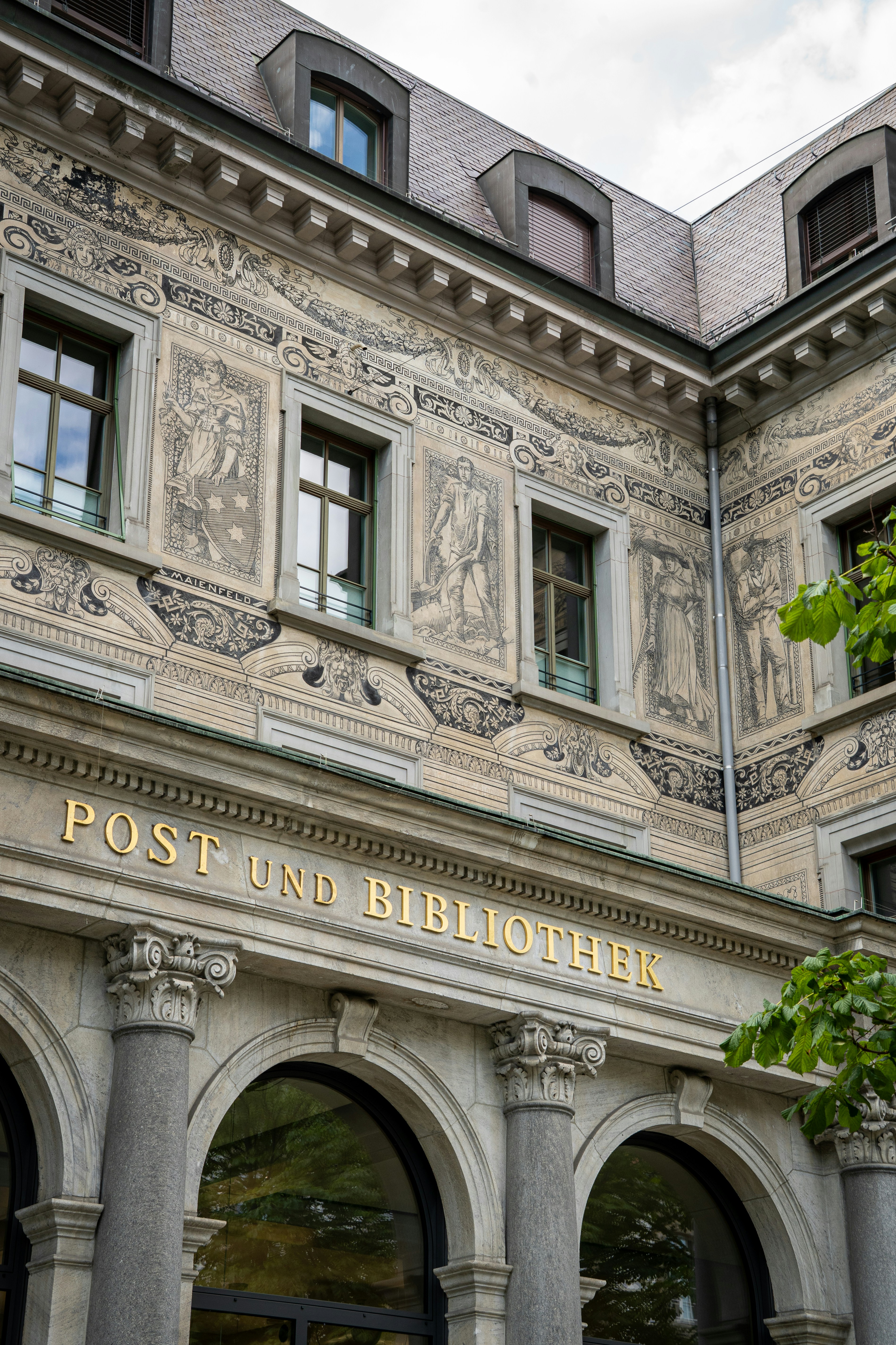 An elegant historic building in Chur, Switzerland, with the golden inscription "Post und Bibliothek" above grand stone arches. Its upper façade is richly decorated with intricate sgraffito artwork depicting classical figures, reflecting the city’s blend of culture, history, and architectural finesse.
