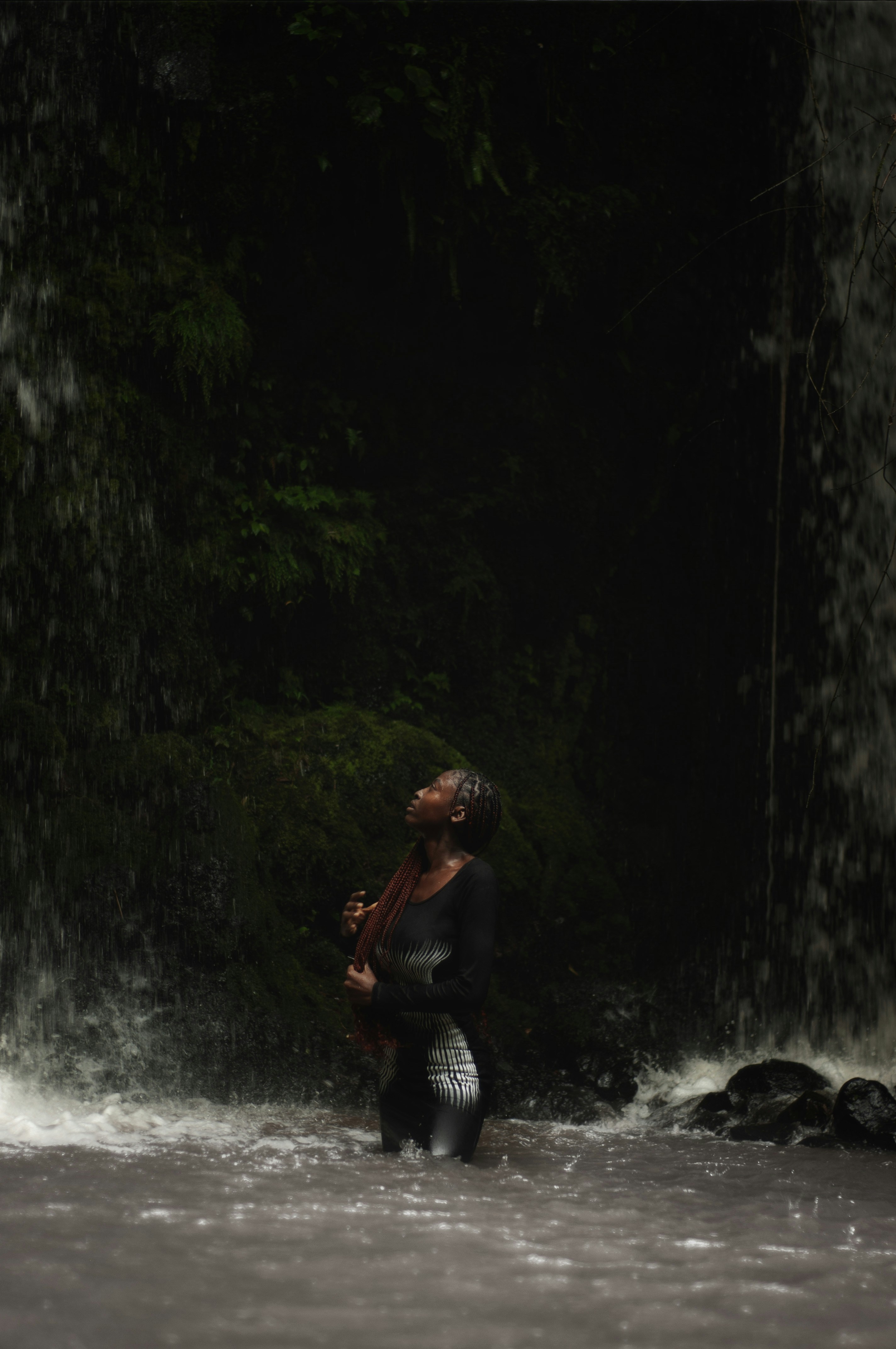 Woman stands beneath a waterfall, looking upwards.