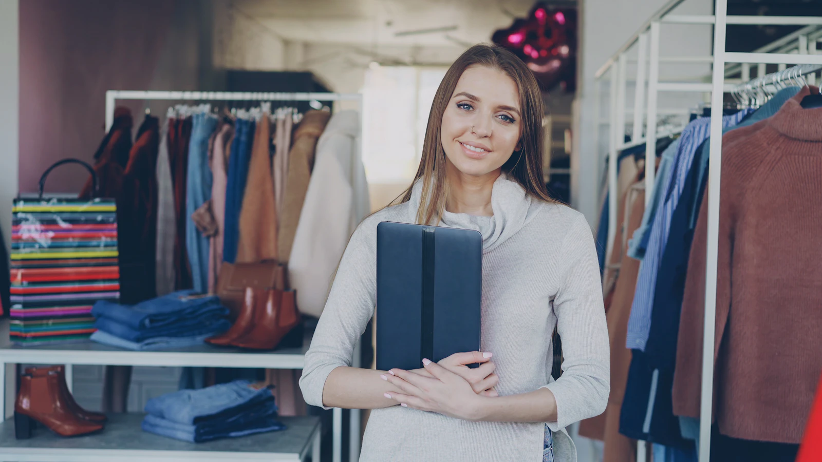 A young boutique owner standing in her clothing store, holding a tablet and smiling at the camera, with racks of clothing behind her