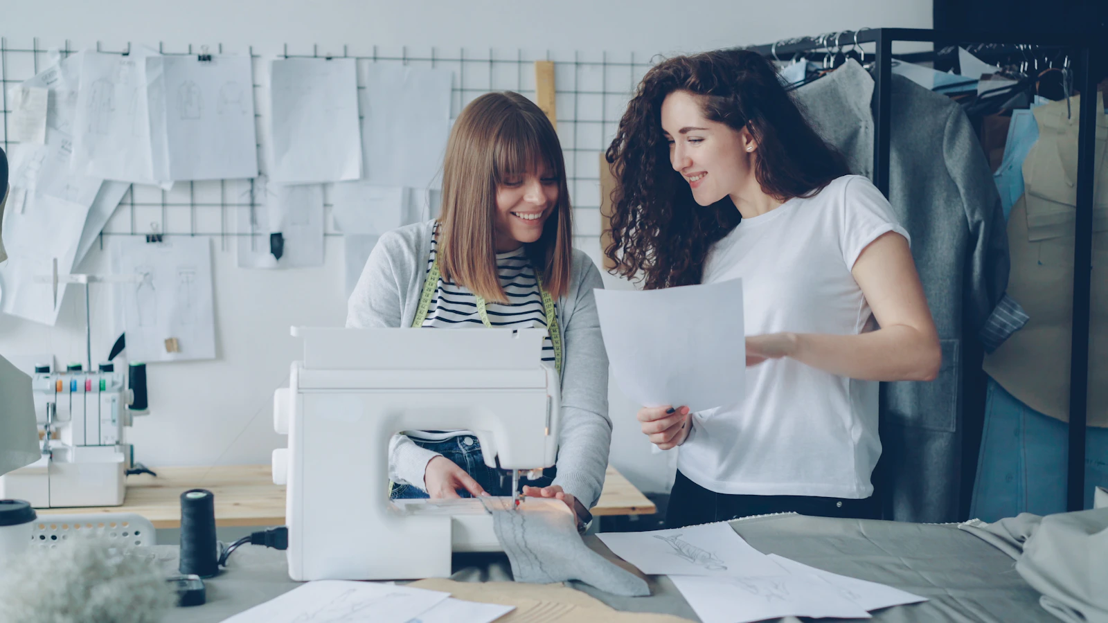 Two women collaborating in a fashion studio, one operating a sewing machine while the other reviews design sketches.