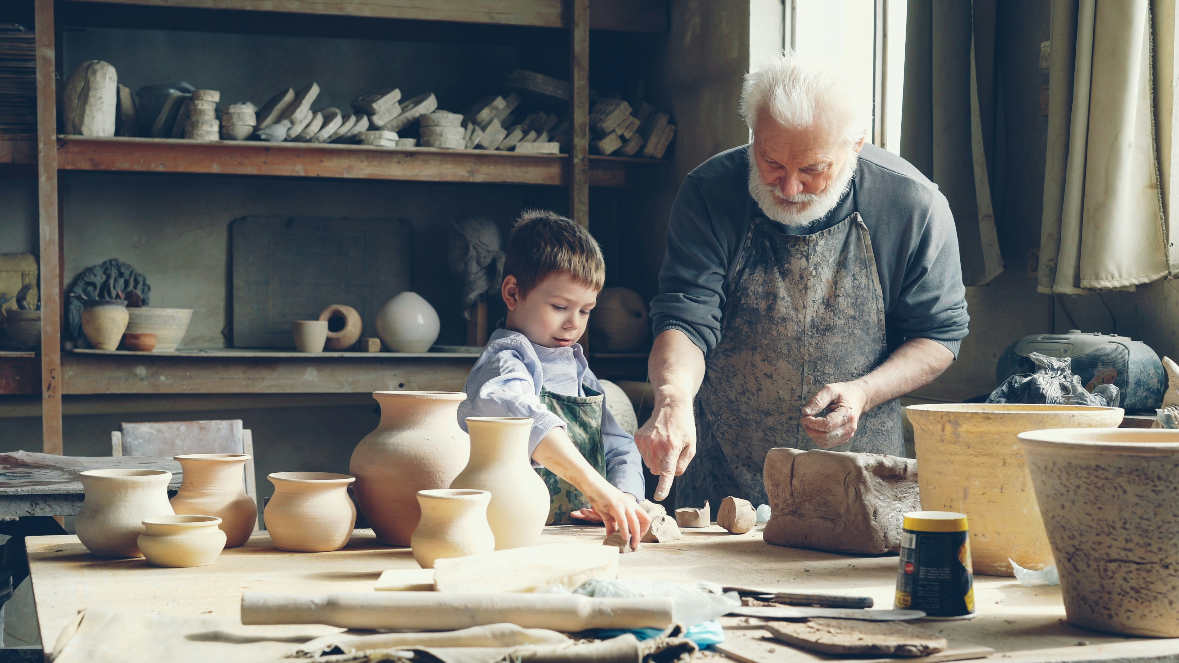 Cheerful boy helping grandfather in potter's workshop