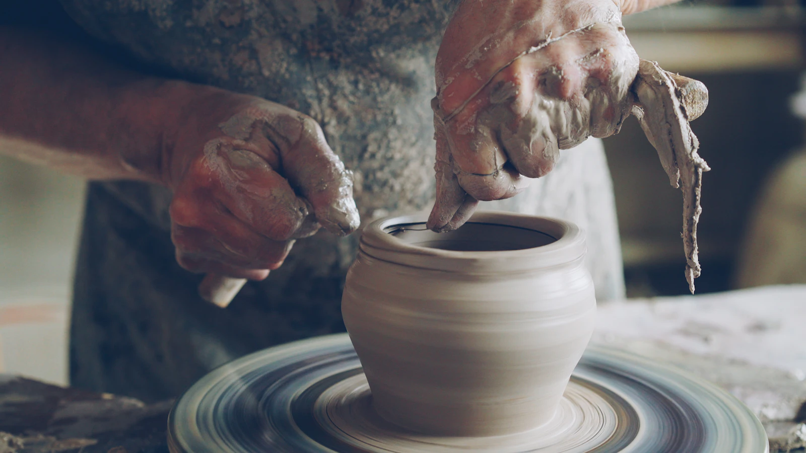 A potter's clay-covered hands shaping a vessel on a spinning pottery wheel
