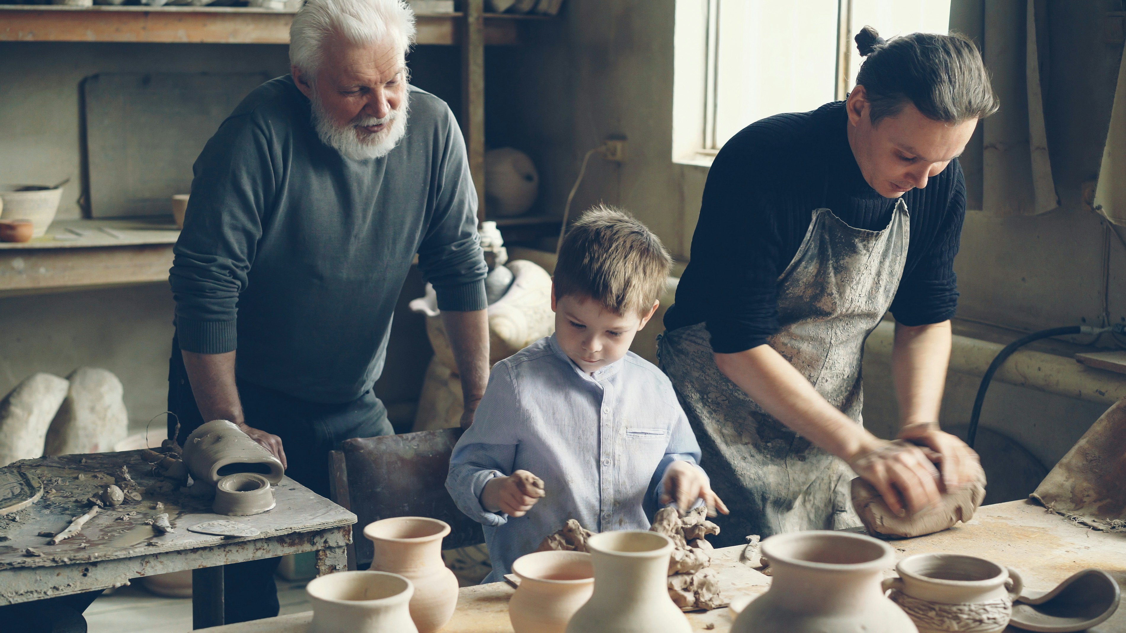 Professional male potter is kneading clay on worktable in home studio while his son is helping him and his elderly father watching them from behind. Small family business concept.