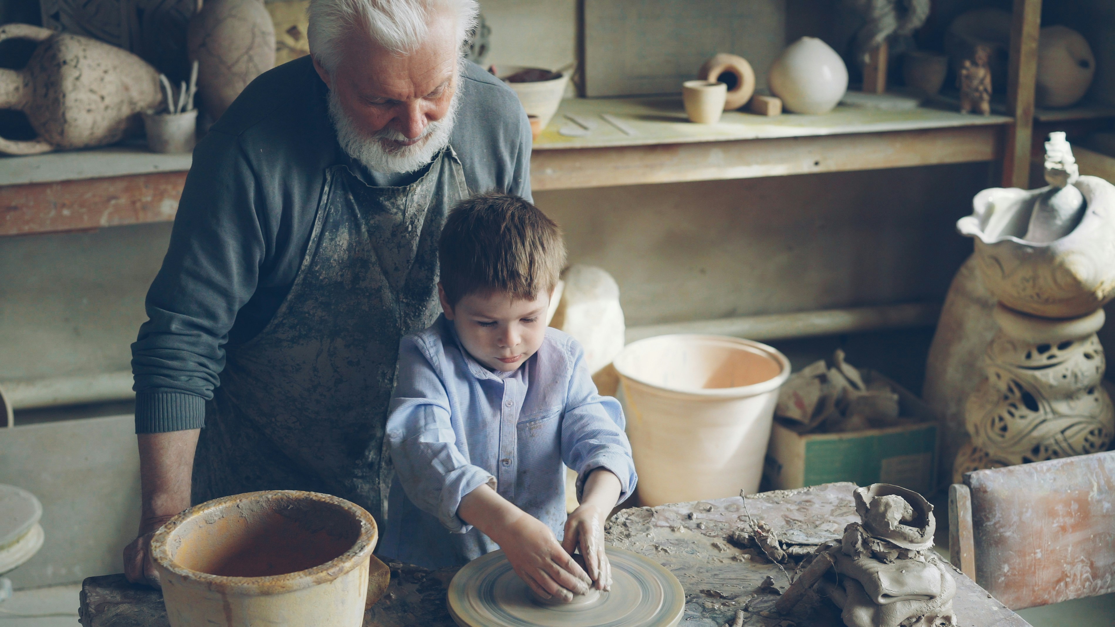 Young cute boy is making clay figure on throwing wheel while his caring grandfather experienced sculptor is standing near worktable and watching carefully. Family tradition concept.