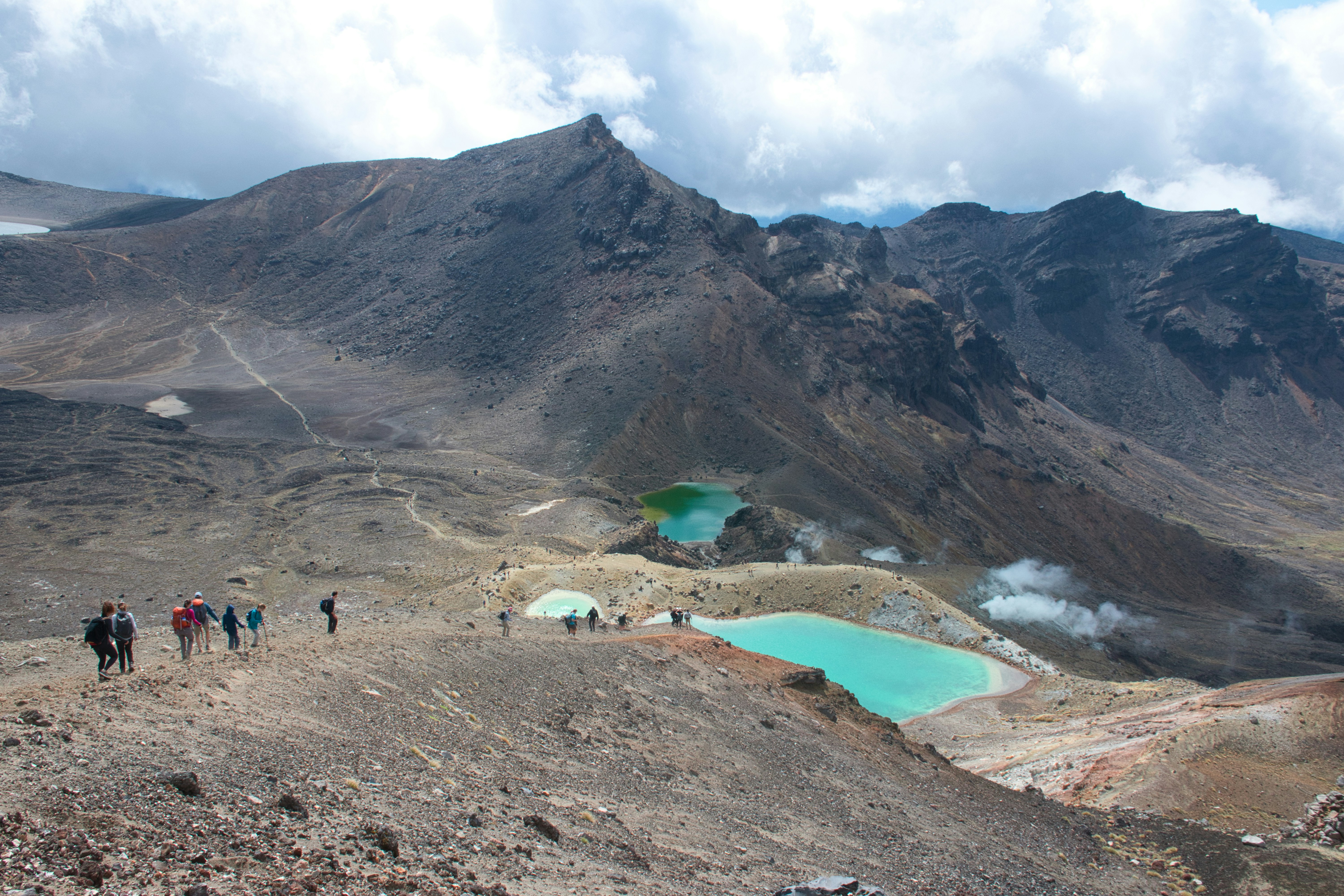 Hikers traverse volcanic terrain near turquoise lakes.