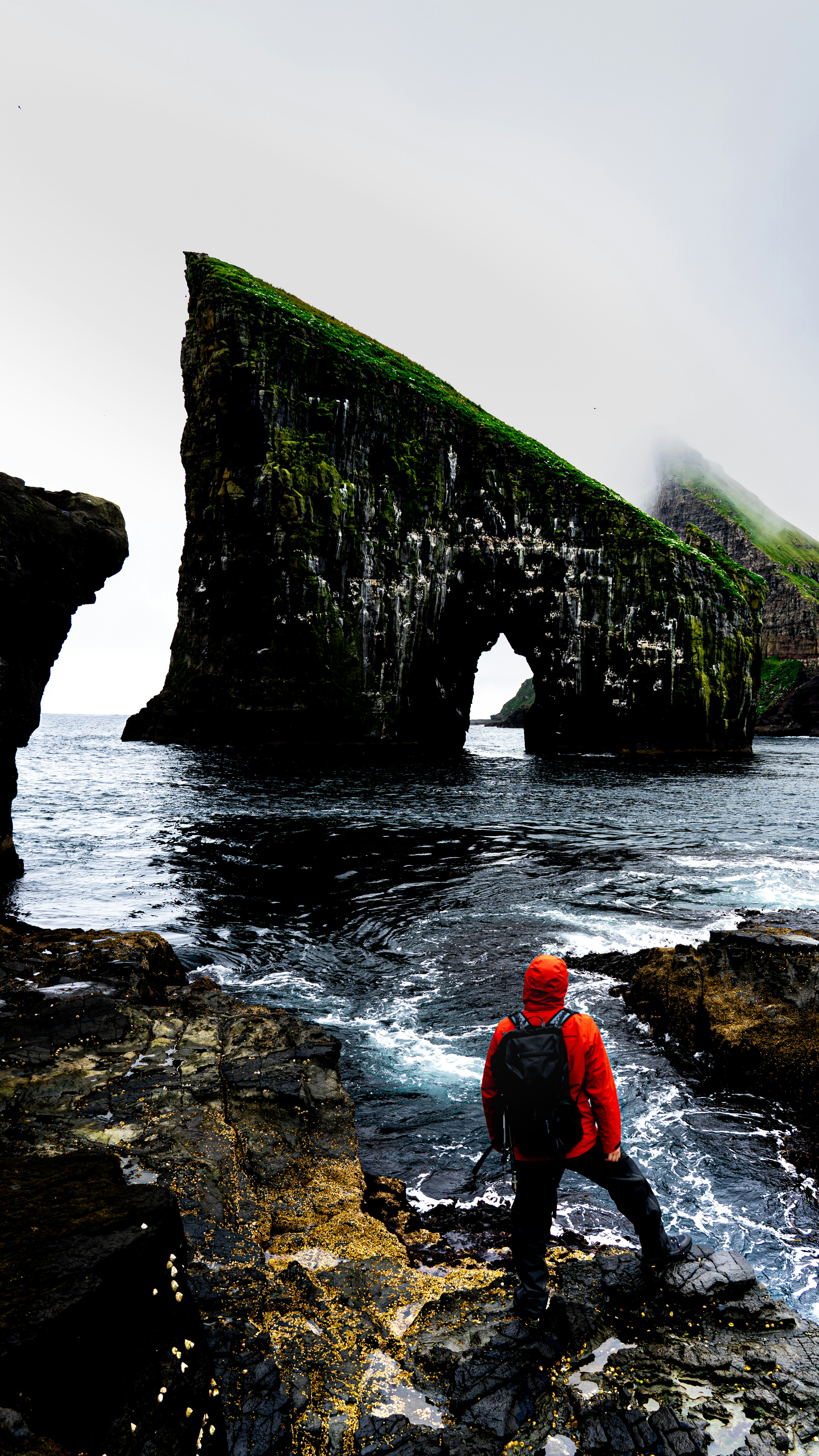 Adventurer gazes at a sea arch in a misty landscape.