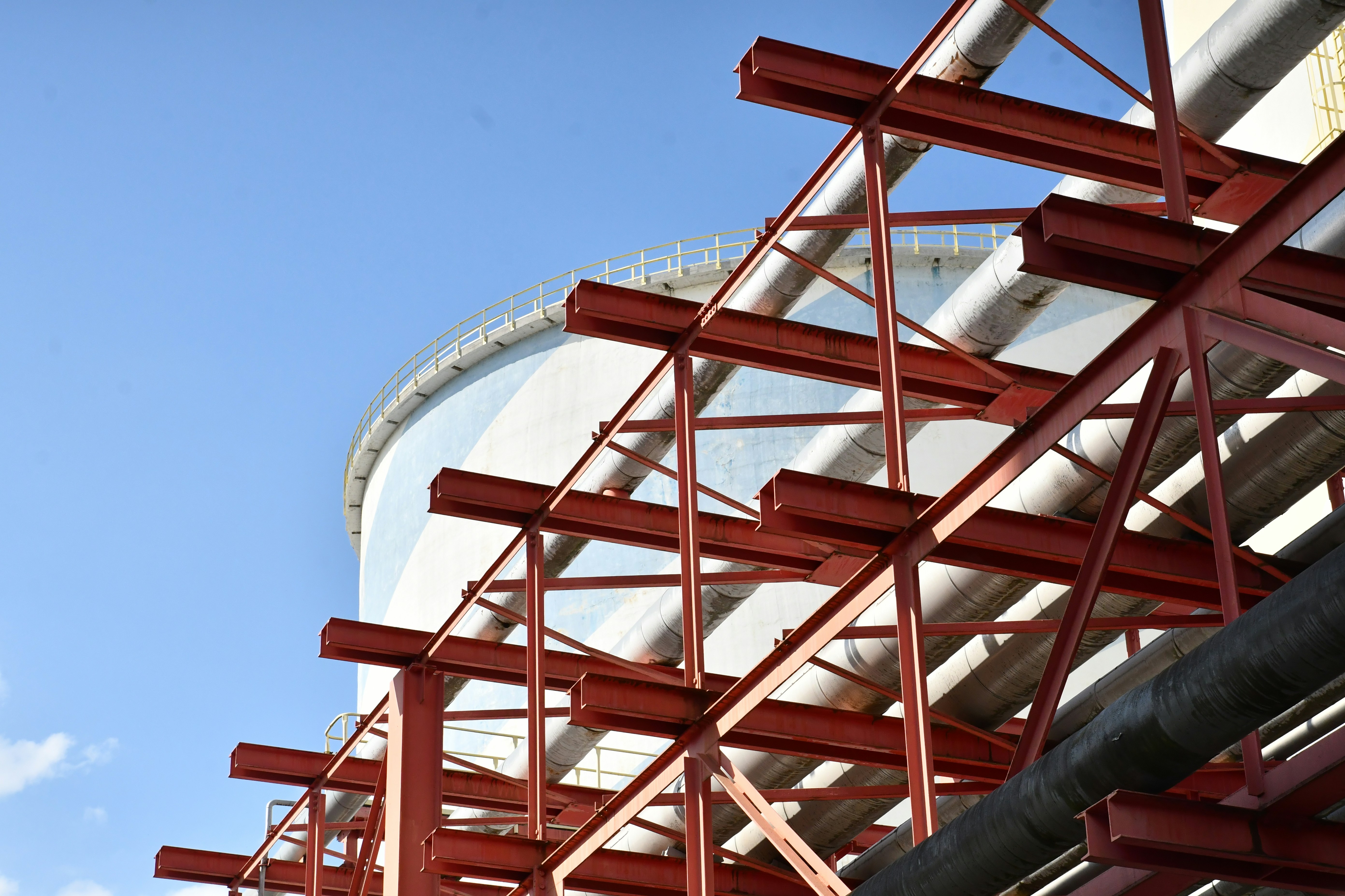Red metal beams support pipes near a large tank.