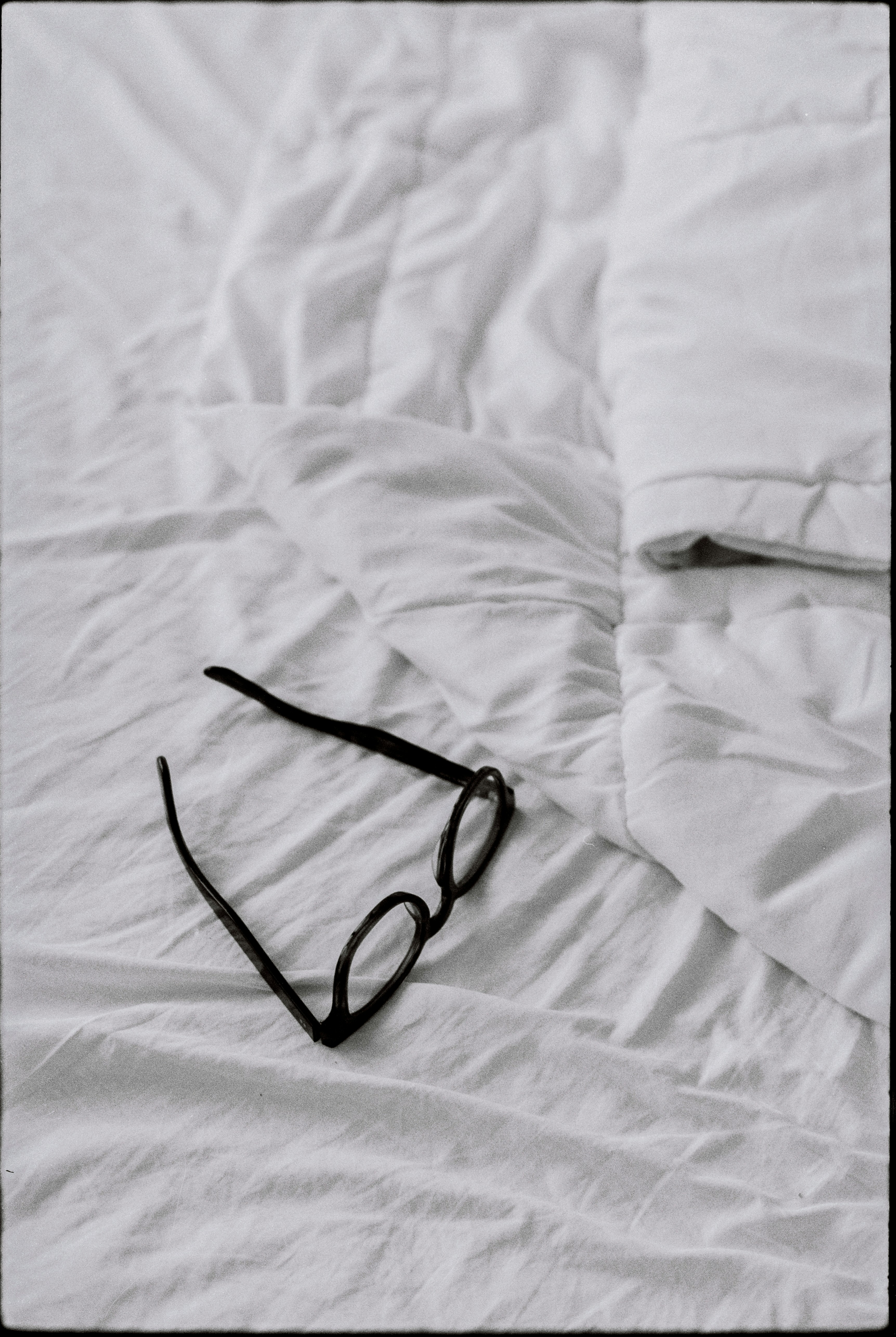 Eyeglasses rest upon a rumpled, white bed.
