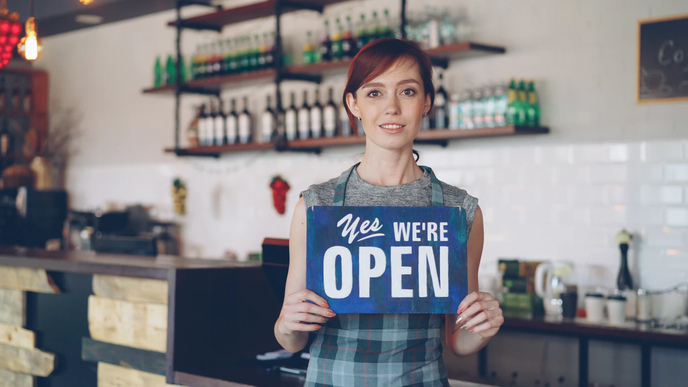 A smiling woman in an apron holds a “Yes we’re open” sign behind a café counter
