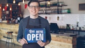 A shopkeeper is smiling and holding an "open" sign.