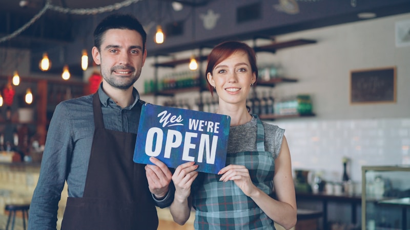 A smiling couple holds an open sign outside their boutique