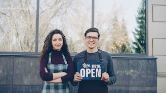 A couple smiles with an "open" sign.