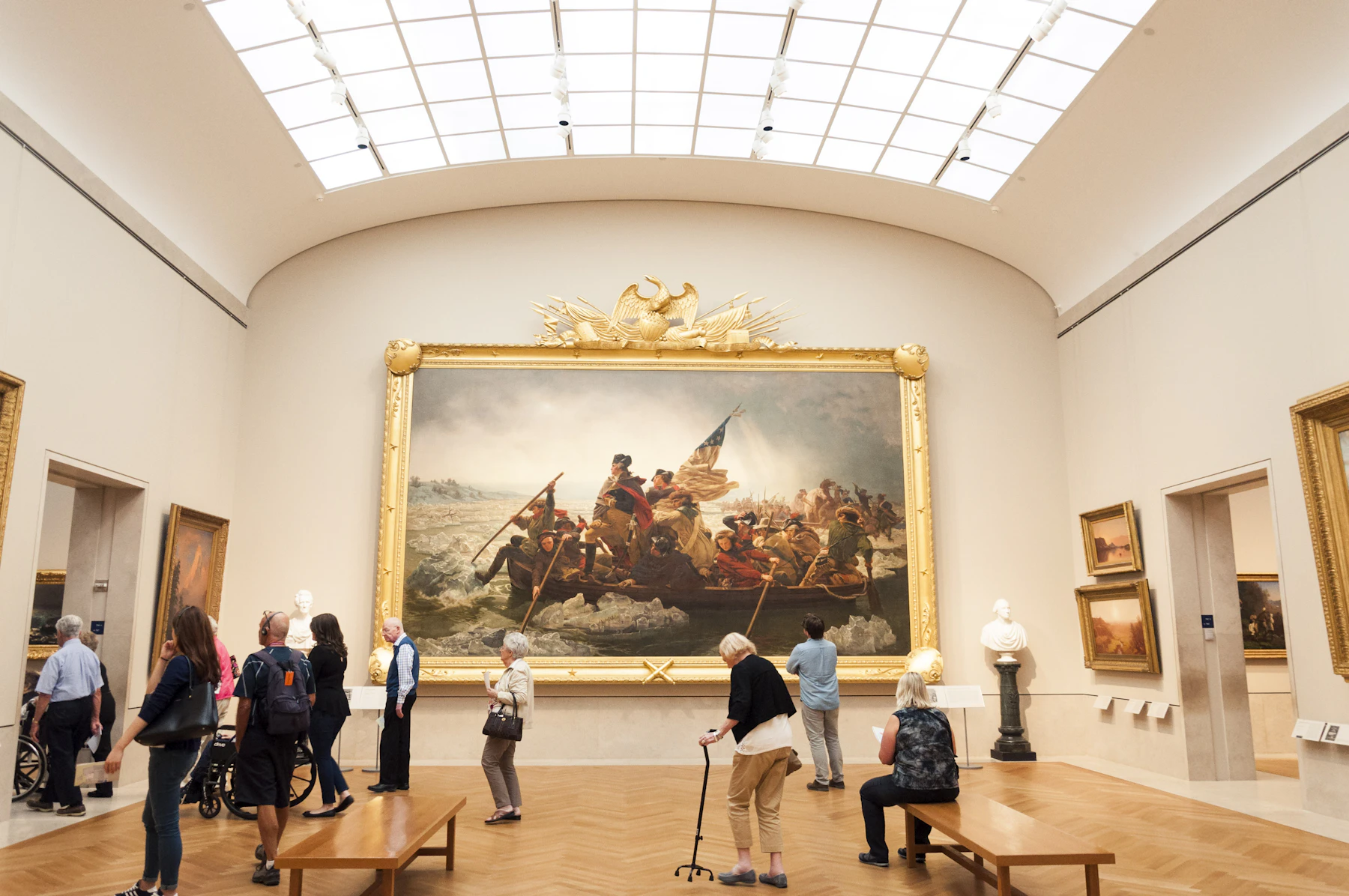 A skylit museum gallery with visitors standing and sitting on benches in front of a very large history painting in a gilt frame