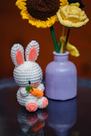 A crochet bunny holds a carrot near flowers.