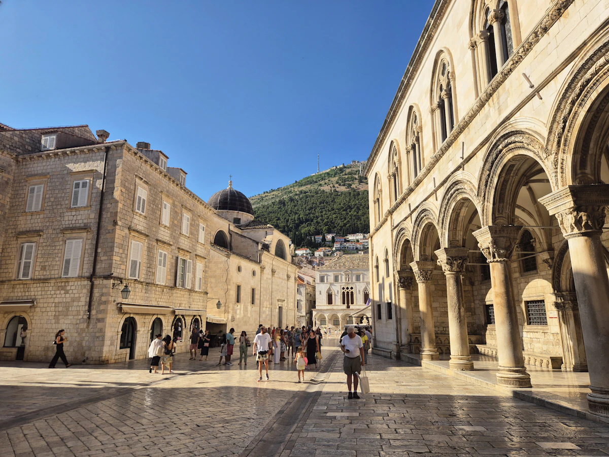 People walking through a sunlit limestone square beside historic arcades in Dubrovnik