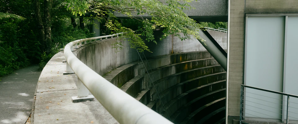 The image depicts a concrete walkway with a curved railing, situated between two buildings. The walkway is bordered by lush green trees and bushes on one side, creating a natural contrast to the man-made structure. On the other side of the walkway, there's an open area with a white building featuring large windows that reflect the surrounding greenery.