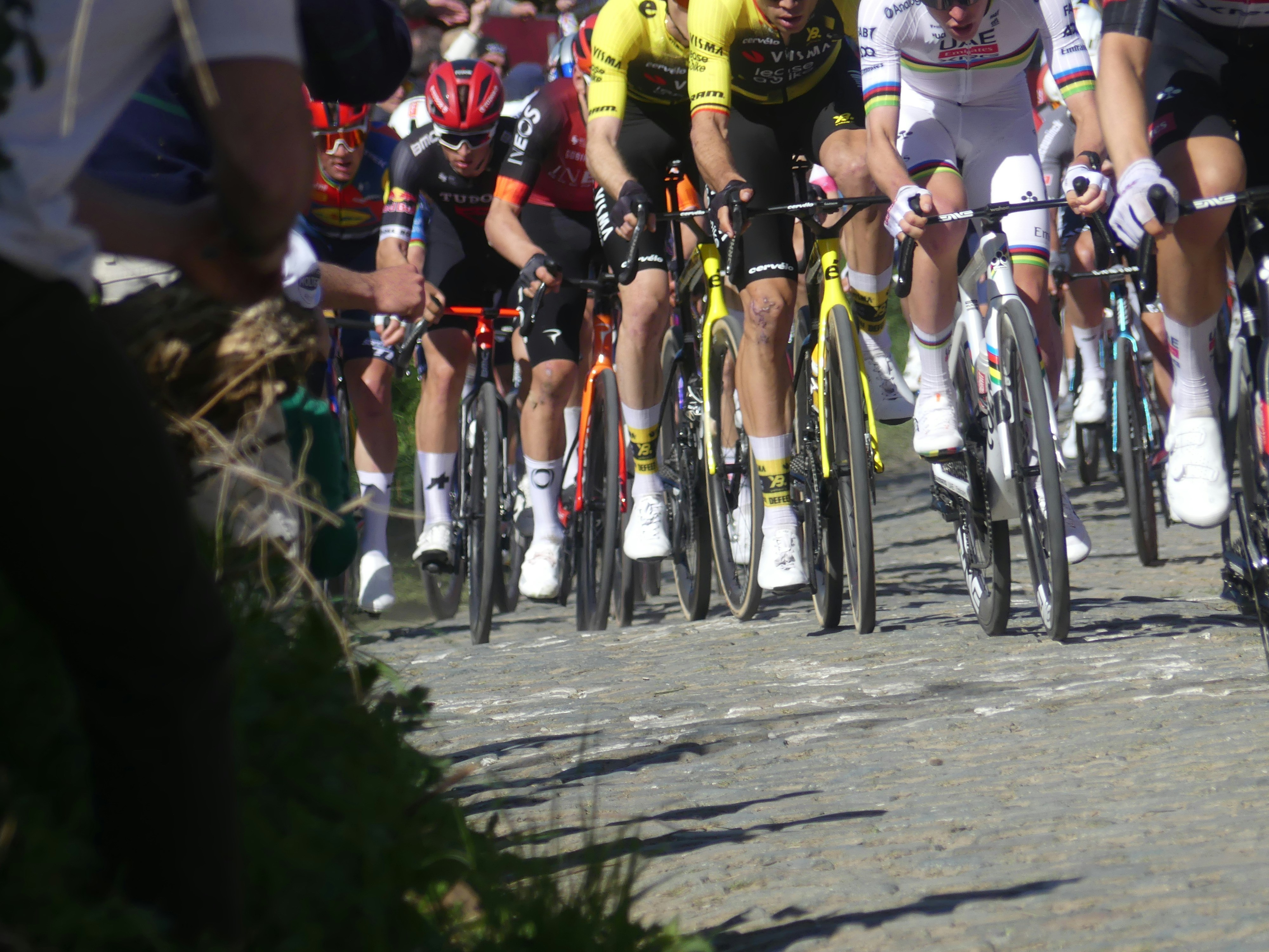 Cyclists race on a cobblestone road under bright sun.