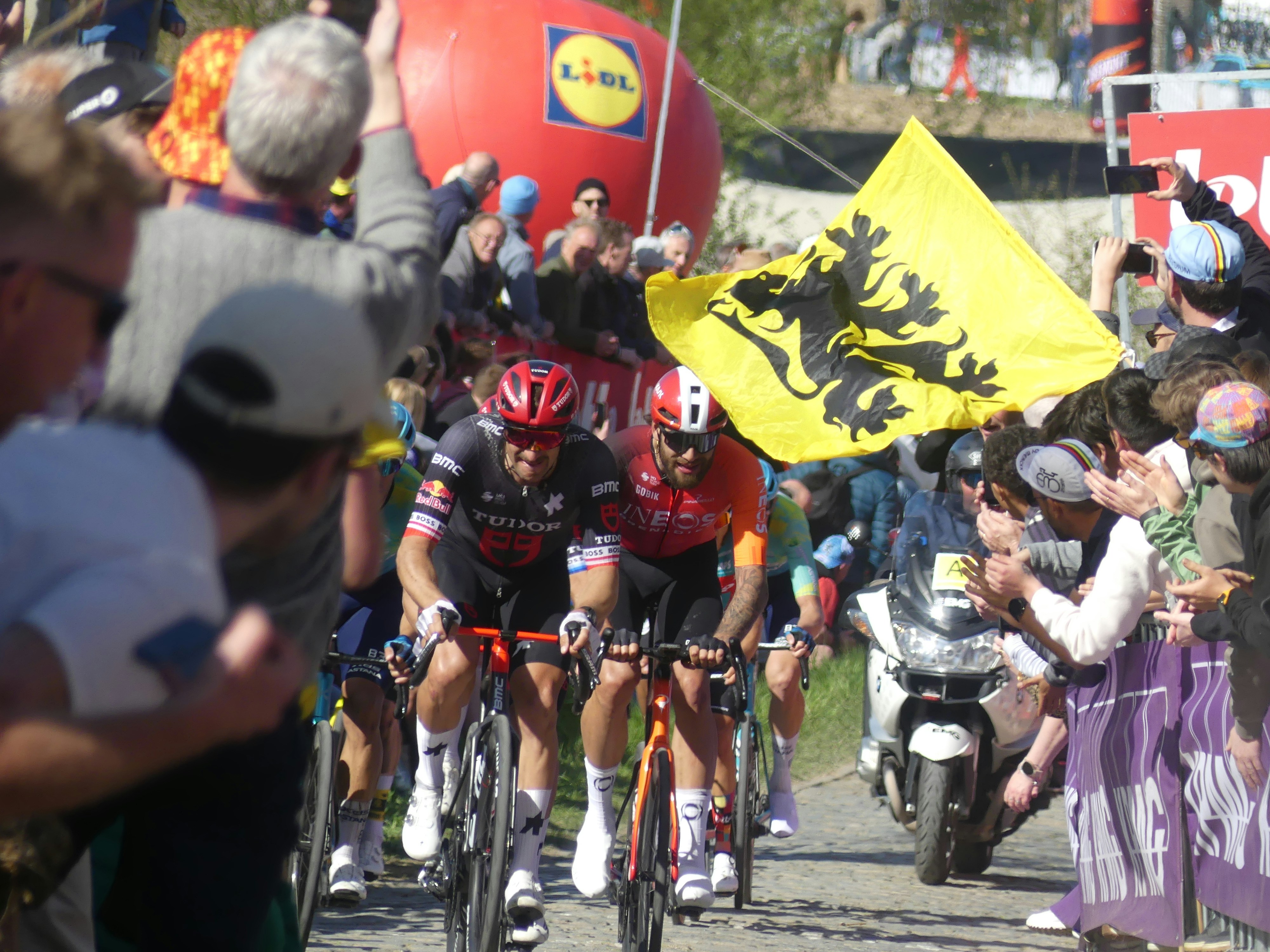 Cyclists race past cheering crowd with flag
