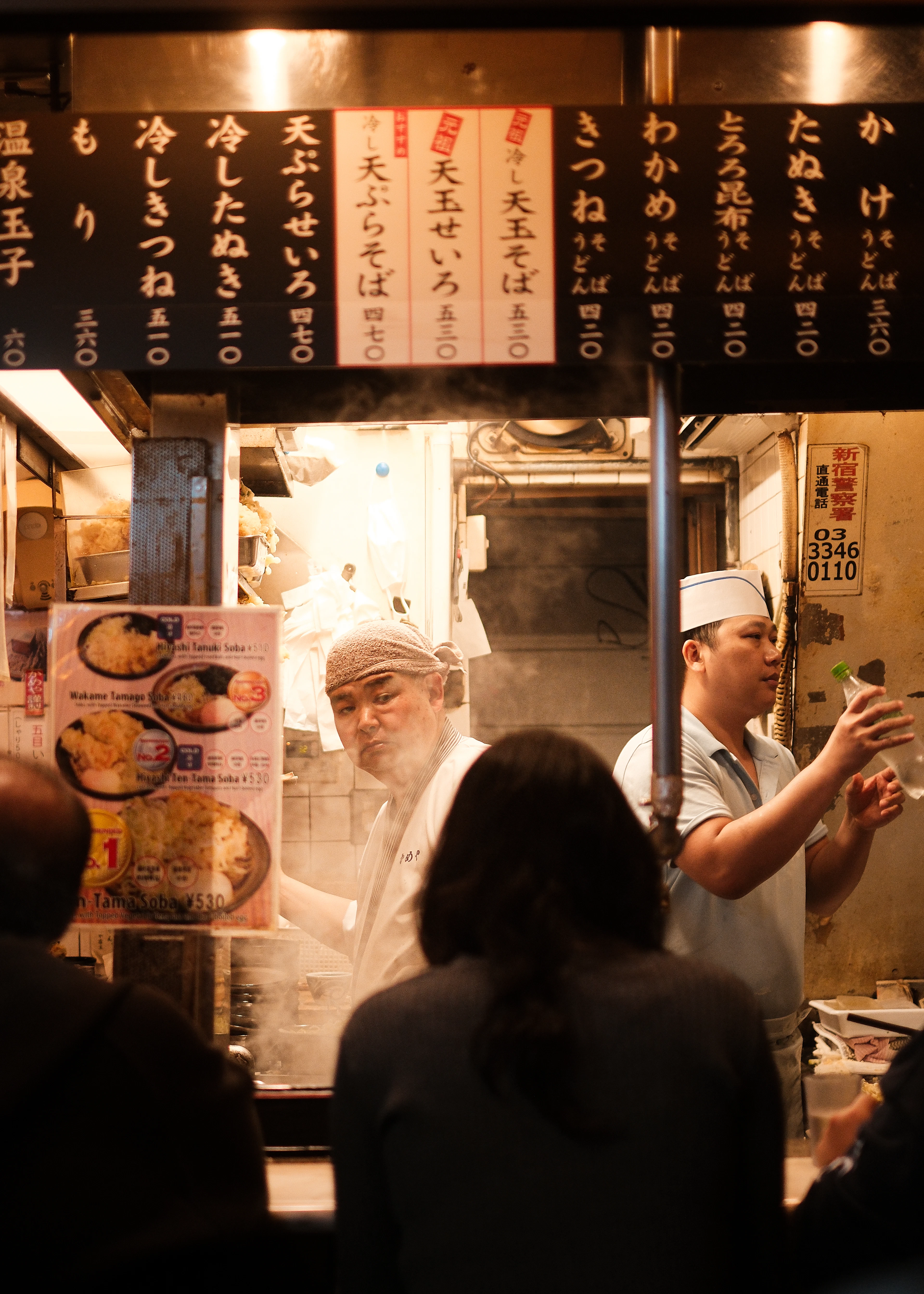 Cooks serving soba and tempura across the counter of a tiny standing noodle bar in Omoide Yokocho, Shinjuku, hand-written Japanese menu boards hanging overhead