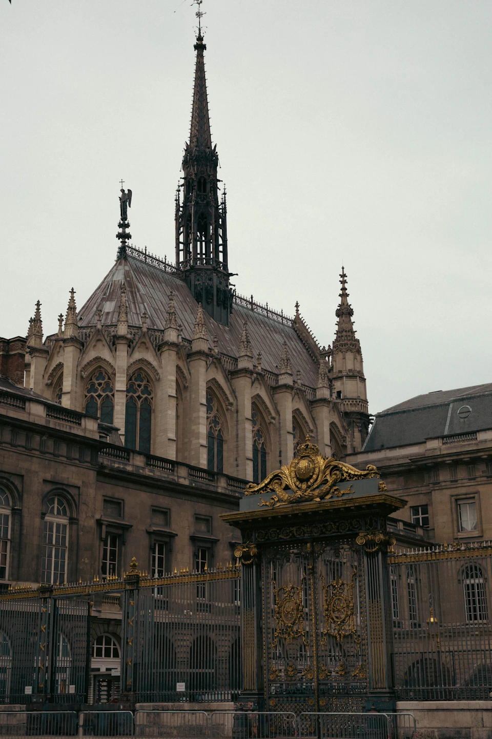 The image depicts a large, ornate building with a tall spire and a clock tower reaching towards the sky. The building is constructed from stone and features intricate carvings on its facade. A gate stands at the entrance to the building, serving as a boundary between the public and private spheres. In front of the building, there are two statues, one of which is a person holding a book, adding an element of historical significance to the scene.

The sky above is overcast, casting a soft light on the scene below. The image does not contain any discernible text or countable objects, and the relative positions of the elements suggest that they are all part of the same architectural ensemble.