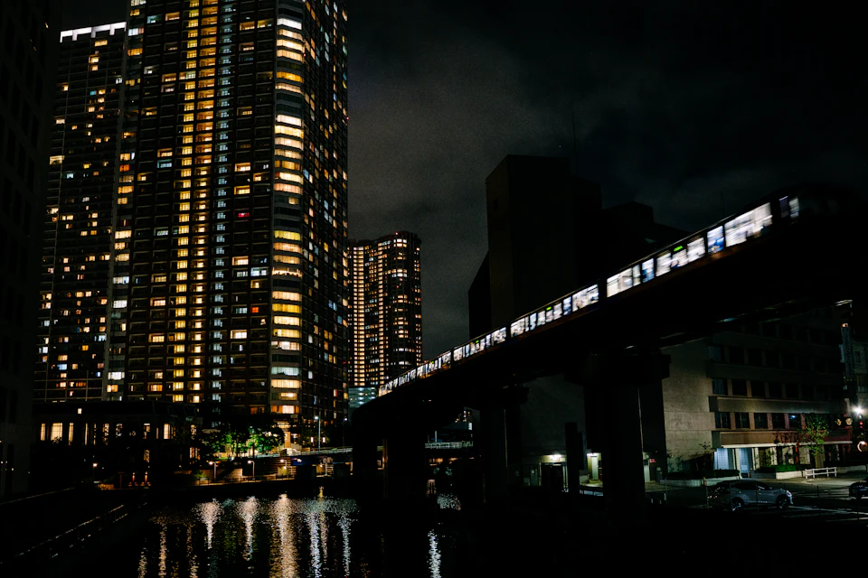 The image depicts a cityscape at night, with a train crossing over a river on an elevated track. The train is illuminated by white lights and appears to be moving from left to right across the frame. In the foreground, there are tall buildings that cast long shadows onto the water below. The sky above is dark, creating a contrast between the bright city lights and the darker night sky.