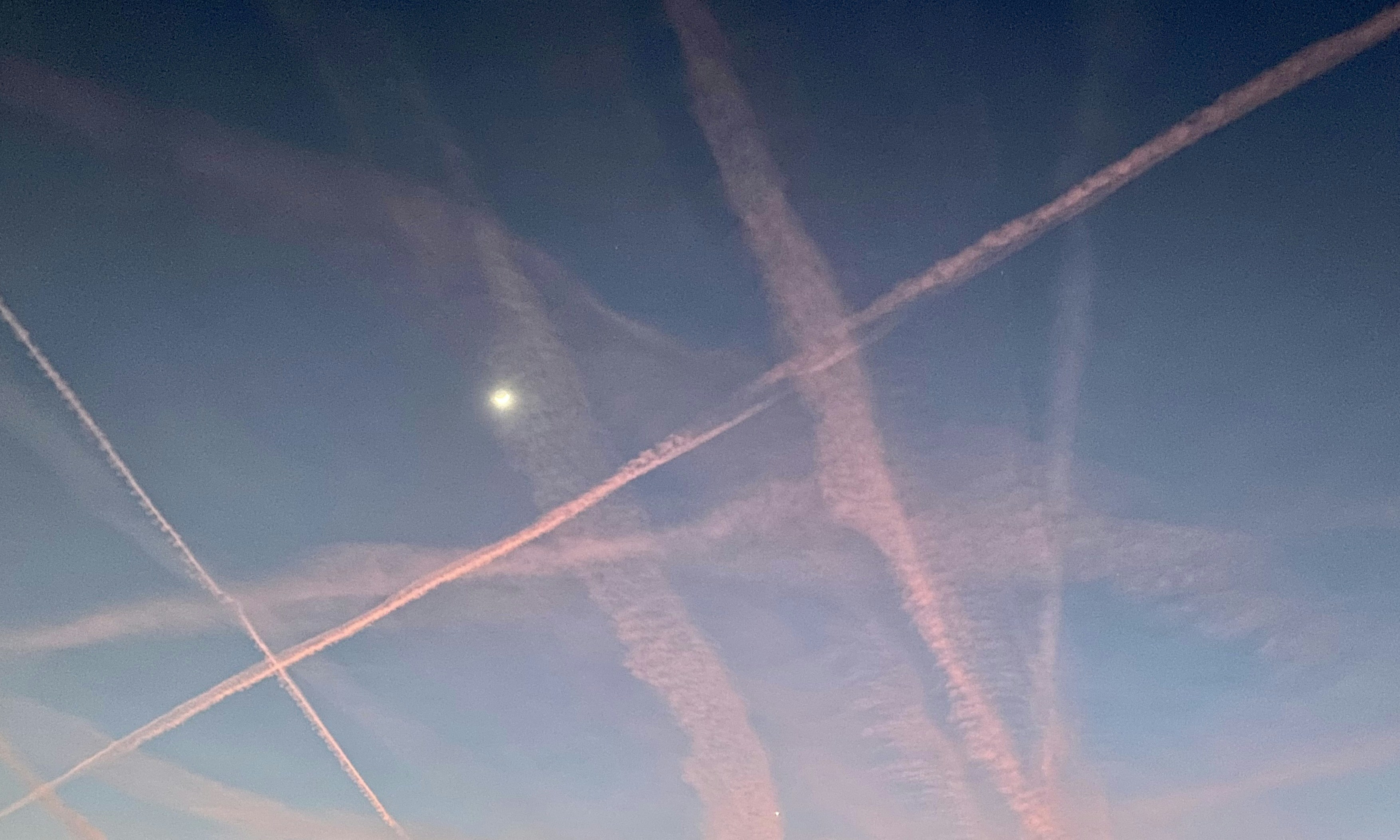 Plane trails crisscross a sky with a moon.